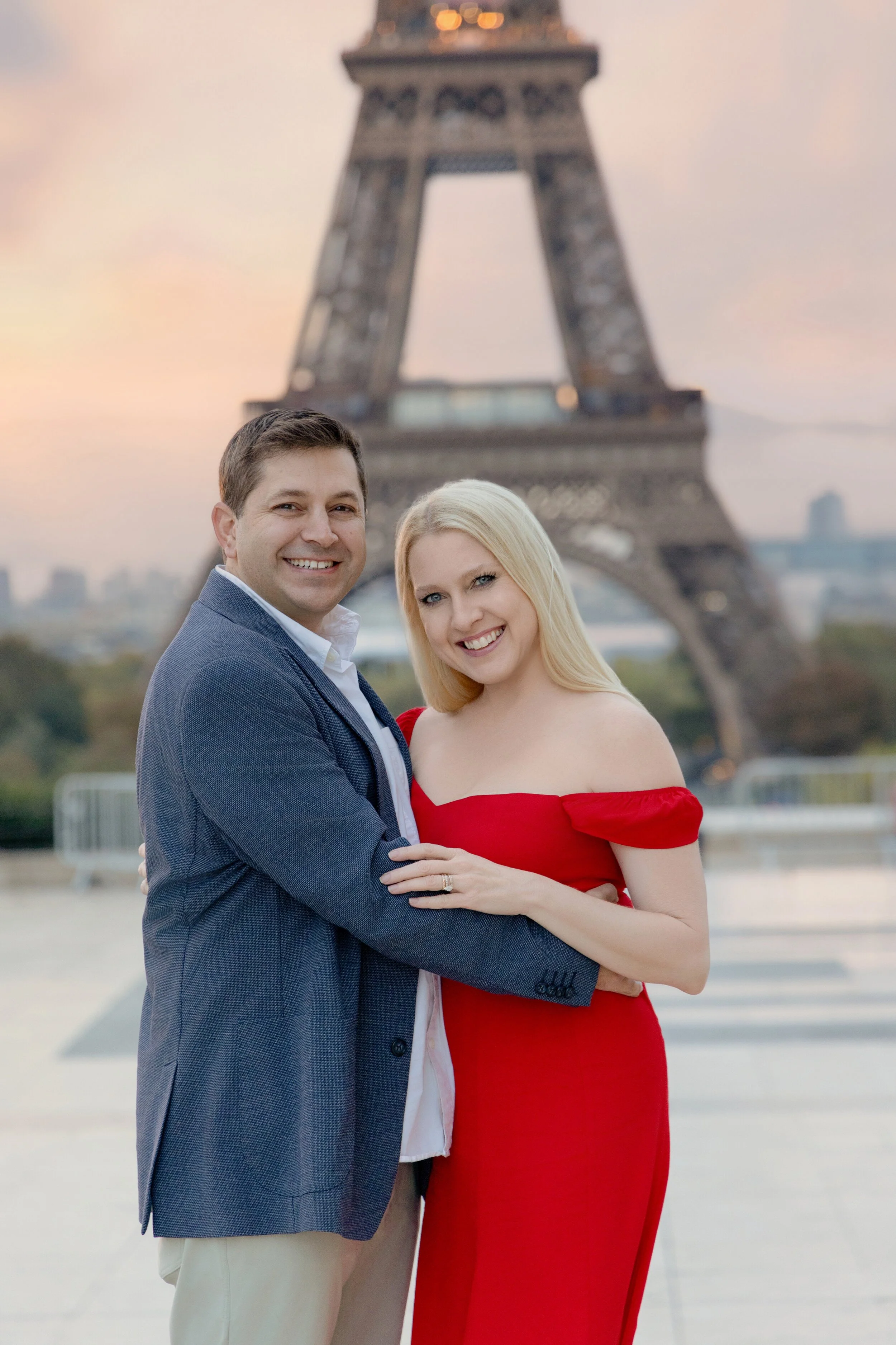 A smiling couple standing in front of the Eiffel Tower in Paris during sunset. The man has short brown hair and is wearing a blue blazer over a white shirt, while the woman has long blonde hair and is dressed in a red off-the-shoulder gown.