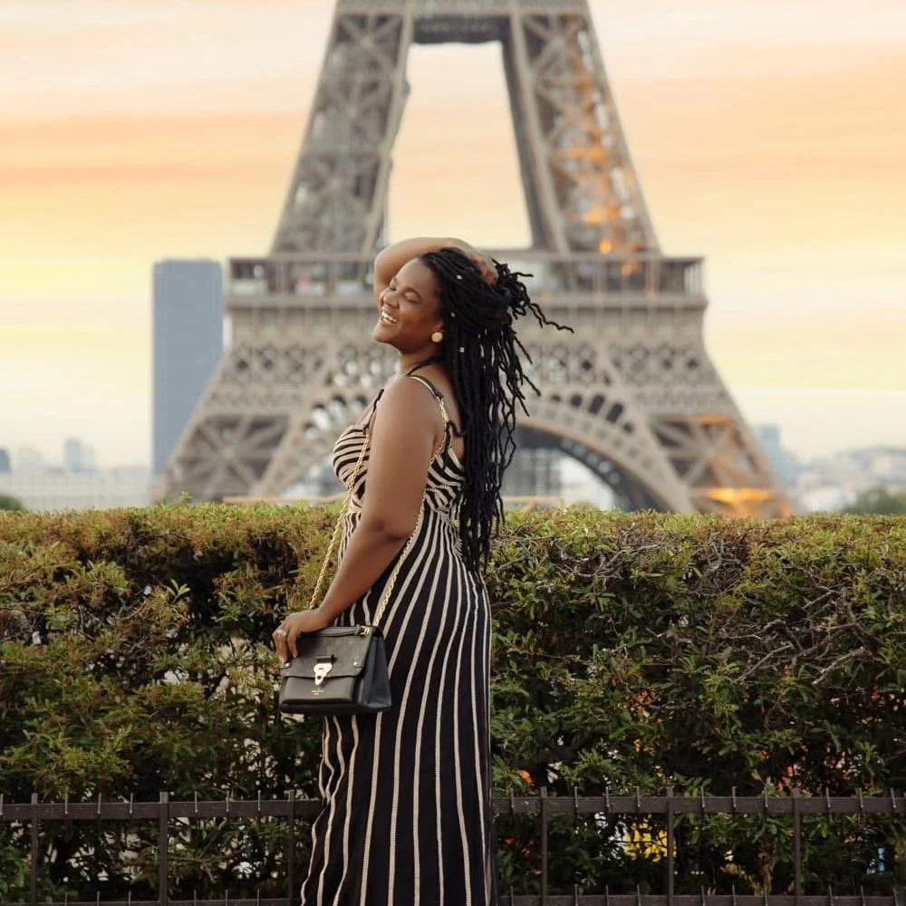 A woman with long dreadlocks smiling and posing in front of the Eiffel Tower at sunset, wearing a striped dress and holding a black purse.