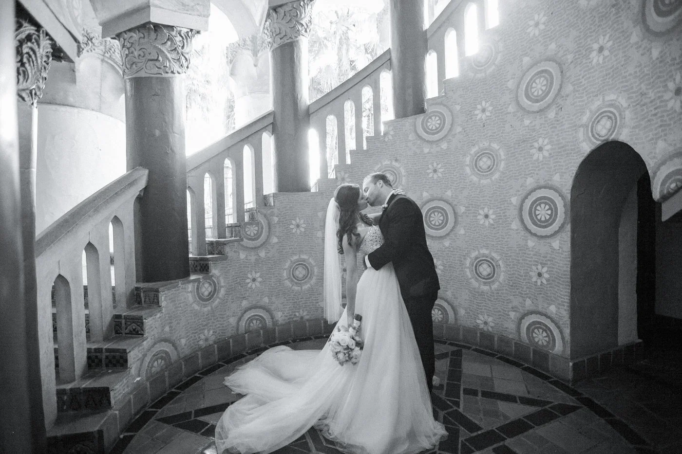 A black-and-white photo of a bride and groom sharing a kiss inside a decorated venue, with ornate columns and patterned walls in the background.