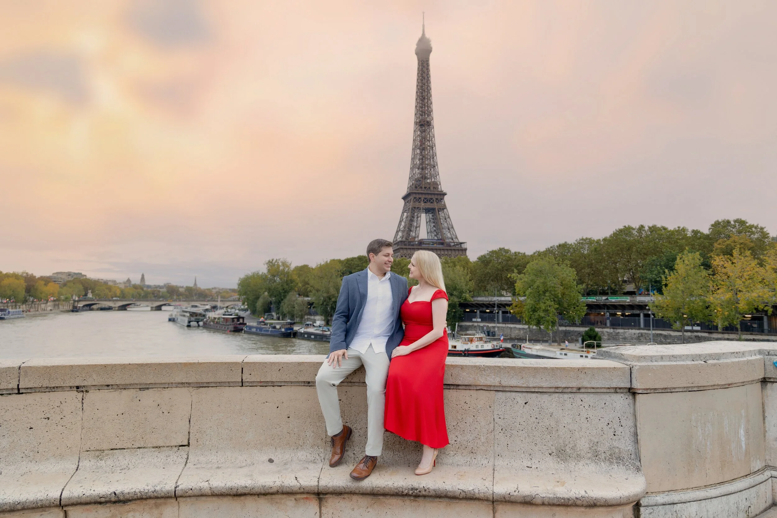 A couple sitting on a stone ledge near the Seine River in Paris, with the Eiffel Tower in the background. The man is wearing a blue blazer, white shirt, and beige pants, while the woman is in a red dress and beige heels. They are looking at each othe