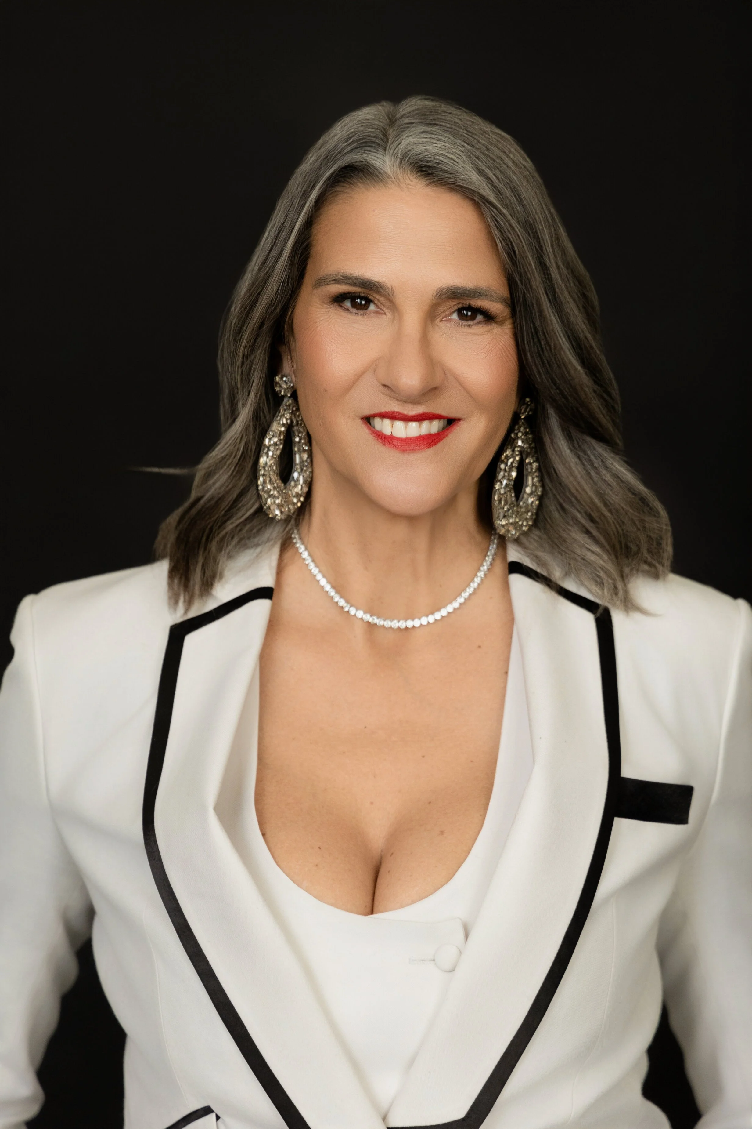 A woman with gray hair wearing a white blazer with black trim, red lipstick, and statement jewelry, smiling at the camera against a black background. Santa Barbara luxury portrait photography