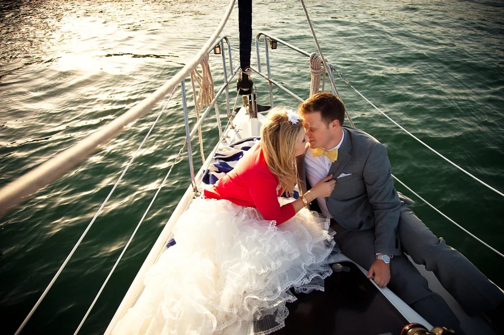A couple dressed in formal attire sharing a kiss on a sailboat at sunset.