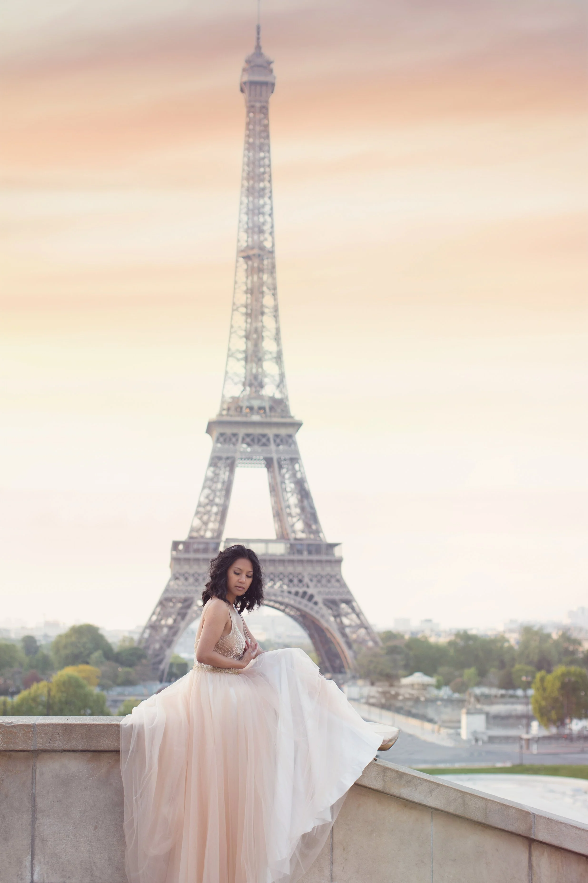 A woman in a flowing beige dress sitting on a ledge with the Eiffel Tower in the background during sunset.