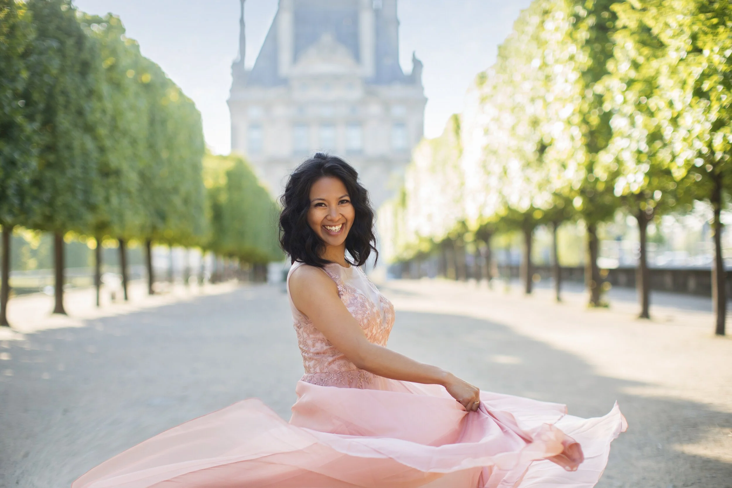 A woman in a pink dress twirling outdoors on a sunlit street lined with green trees, with a historic building in the background. Destination Paris portrait photographer