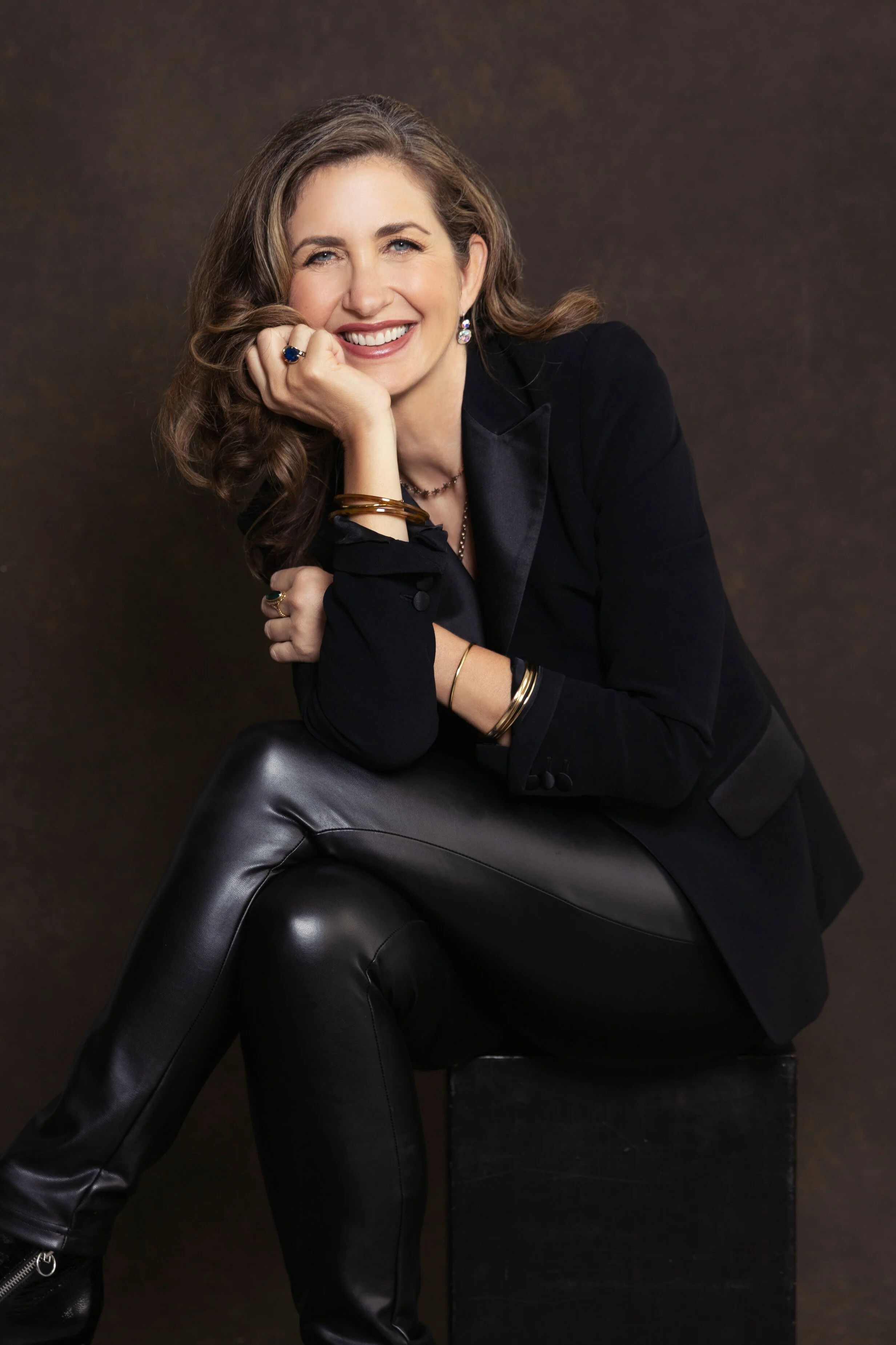 A woman with wavy brown hair smiling, sitting on a black block, wearing a black blazer, black leather pants, and jewelry, posing against a dark background. Santa Barbara luxury portrait photography personal branding