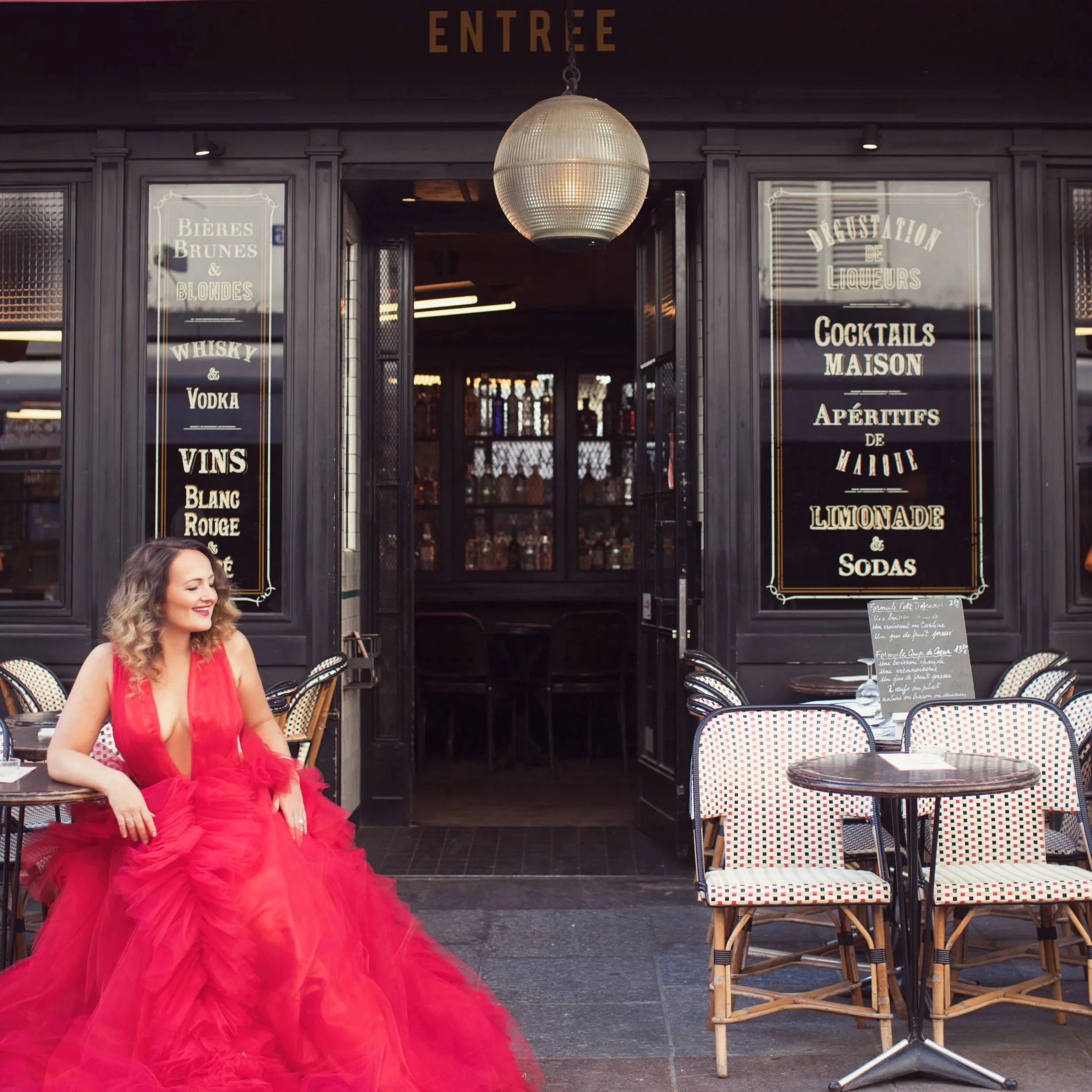 A woman in a red gown sitting outside a French bar or cafe with black storefront, white tables, and chairs. The storefront has gold lettering and menu boards with French words and drink options.