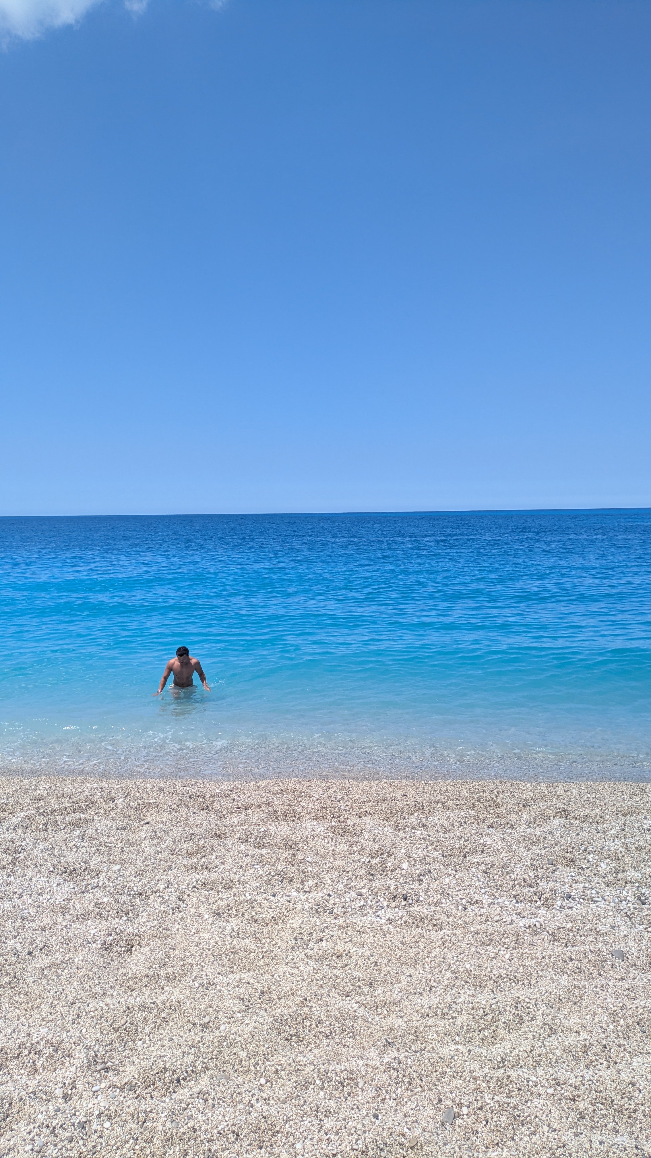 A man in swimming trunks wading in clear blue ocean water near a Milos beach on Lefkada with a bright blue sky above.