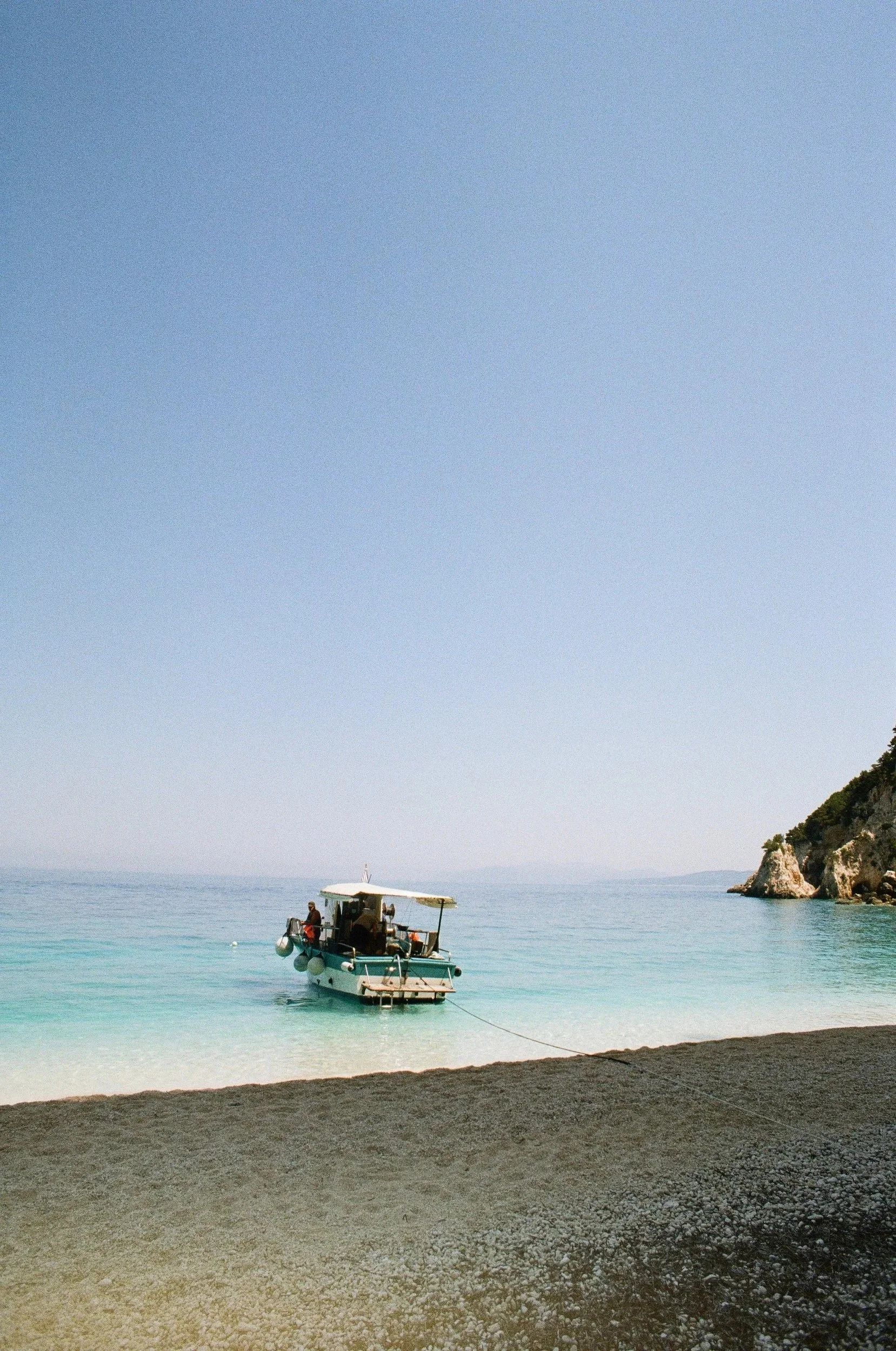 A fishing boat with people on it floating in calm on Kefalonia, turquoise water near a pebble beach with a rocky cliff on the right side and clear blue sky overhead.