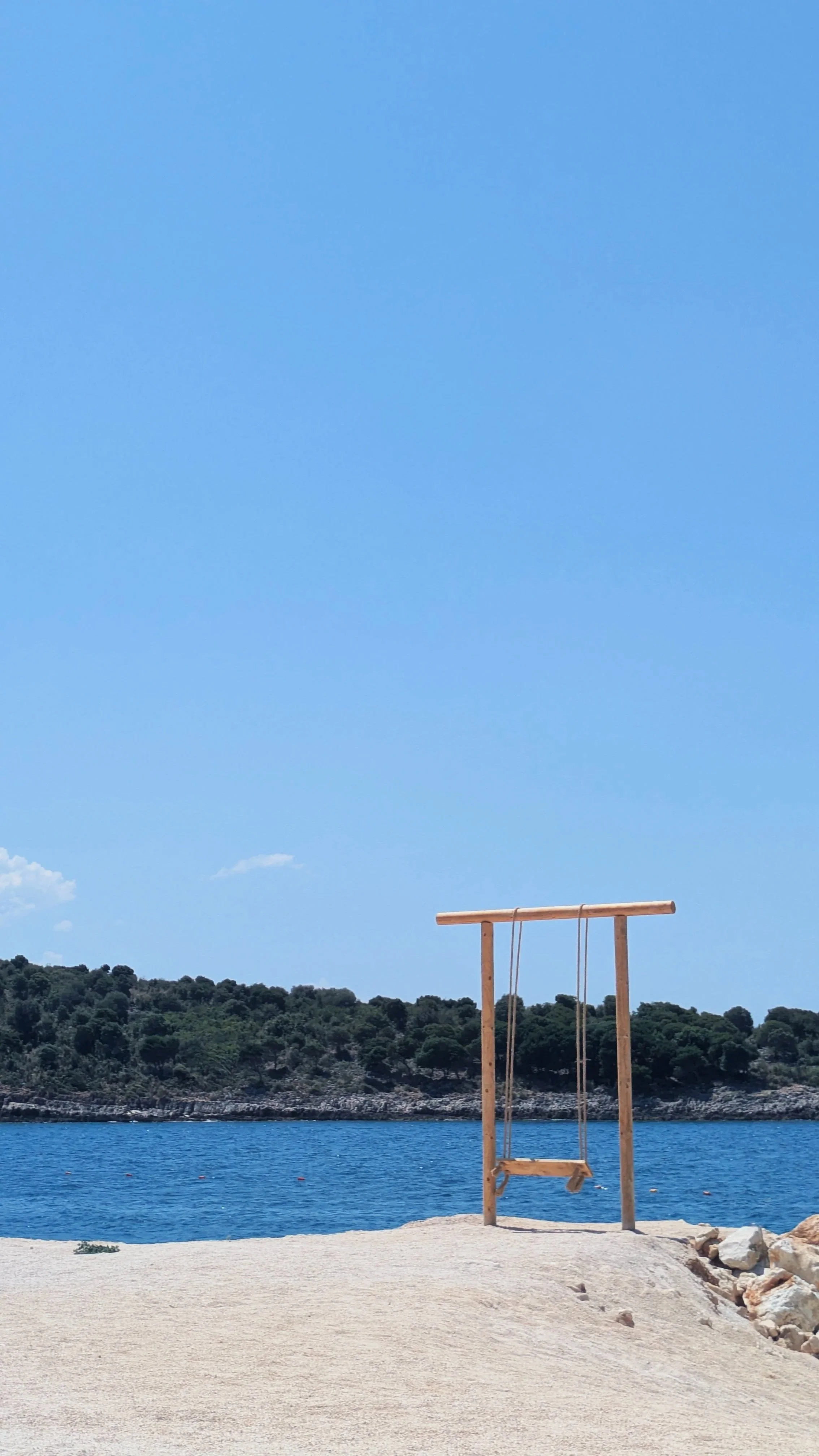 A wooden swing in Sarandë set near the water on a sandy beach with a hilly landscape and trees in the background under a clear blue sky.