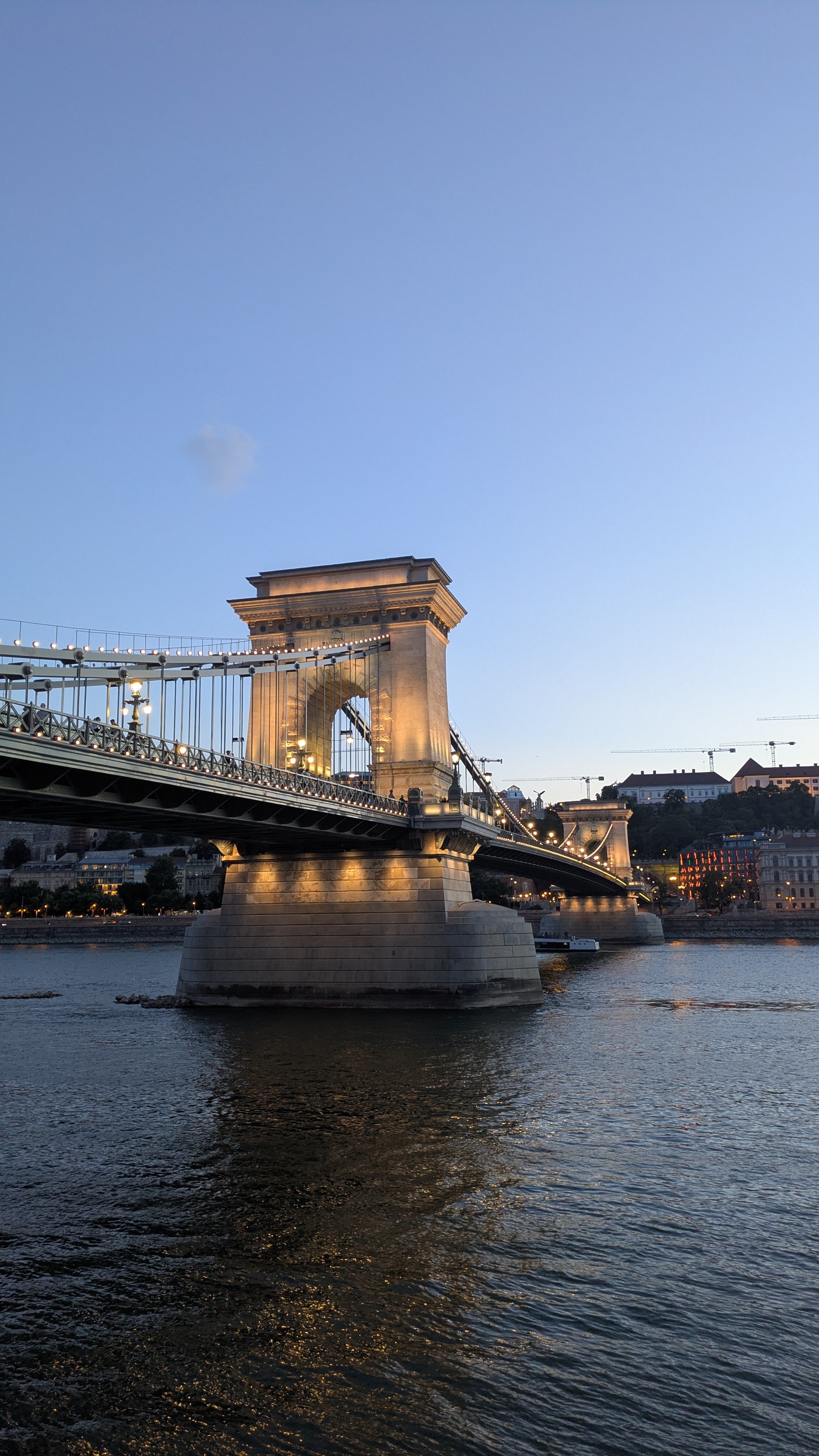 View of the Chain Bridge in Budapest, Hungary, illuminated at dusk with buildings and a hill in the background.