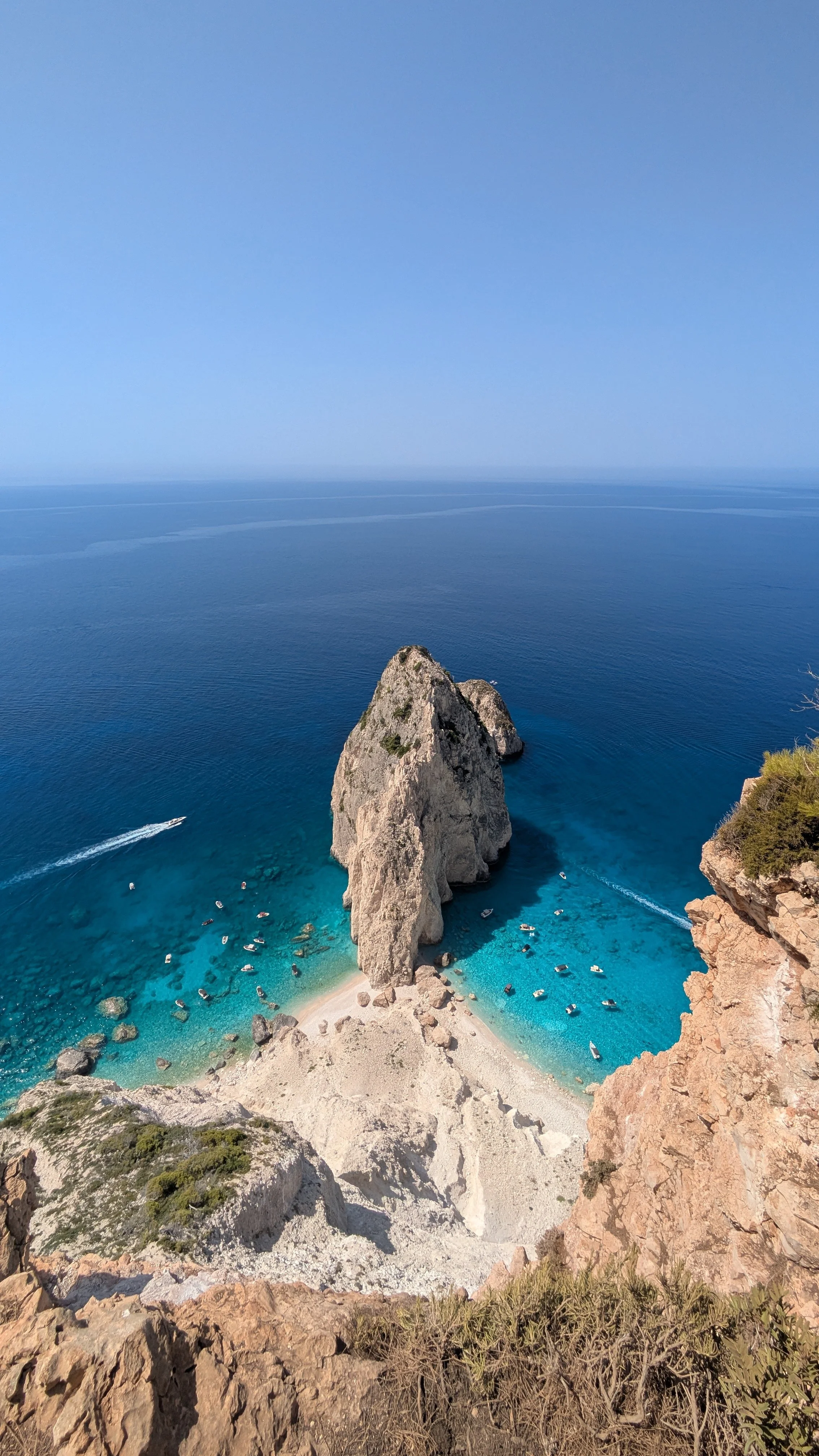 Cliffside view of a large rock formation on Zakynthos in the ocean with boats near the shoreline and clear blue sky.