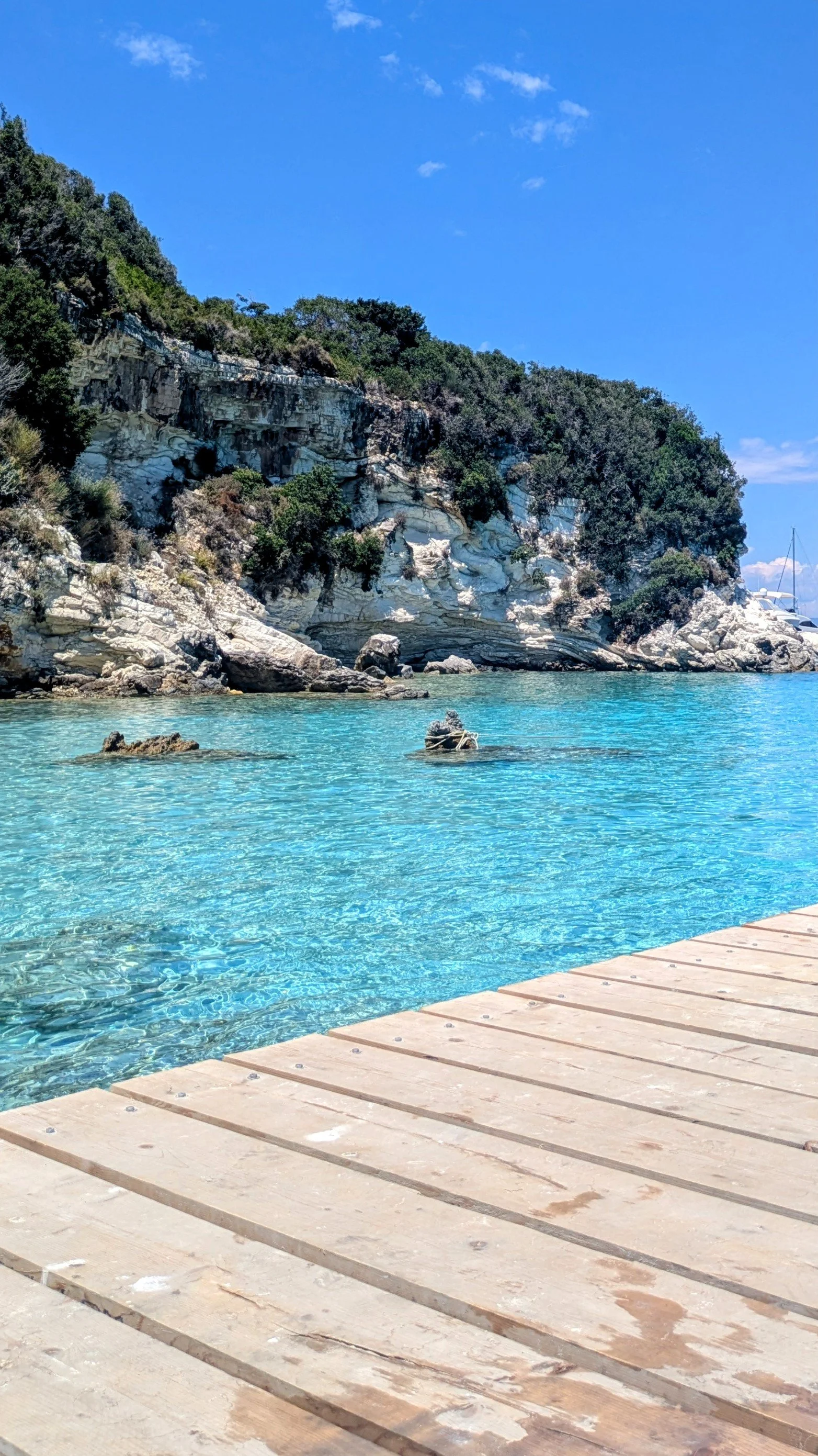 A clear turquoise sea in Antipaxos with a wooden deck in the foreground, rocky cliffs with green vegetation on the side, and a bright blue sky with a few clouds.