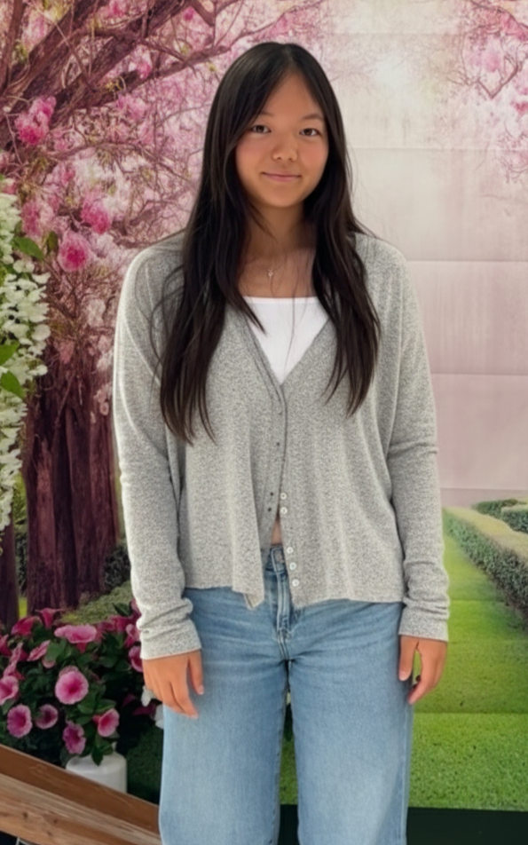 A young woman with long dark hair standing indoors in front of a floral backdrop with pink blossoms and green bushes, wearing a gray cardigan, white top, and jeans.