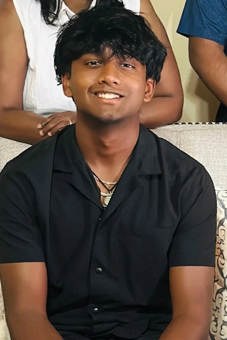 A young man with black, curly hair smiling and sitting on a couch, wearing a black shirt, with two women standing behind him.