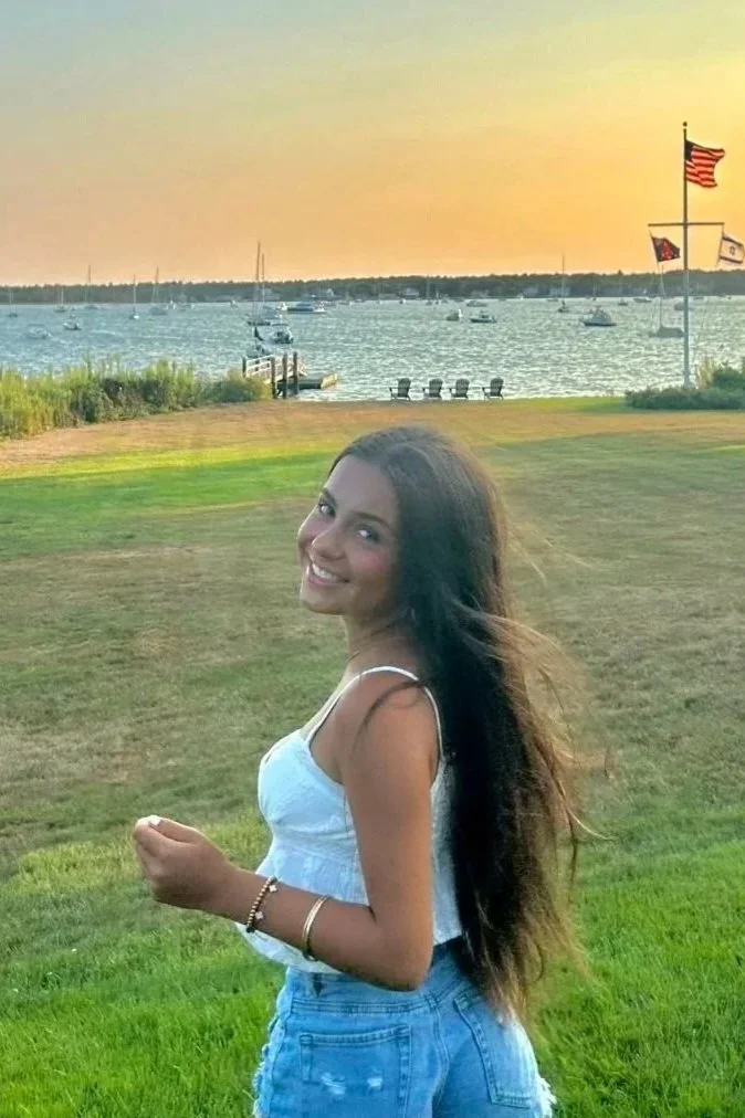 A young woman with long brown hair smiles at the camera while standing on a grassy area near a lake at sunset, with sailboats and American flags in the background.