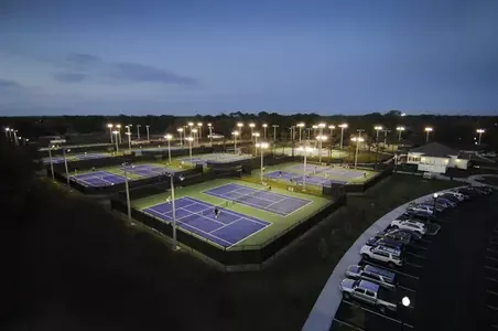 Multiple illuminated tennis courts at dusk, surrounded by parking lots with cars.