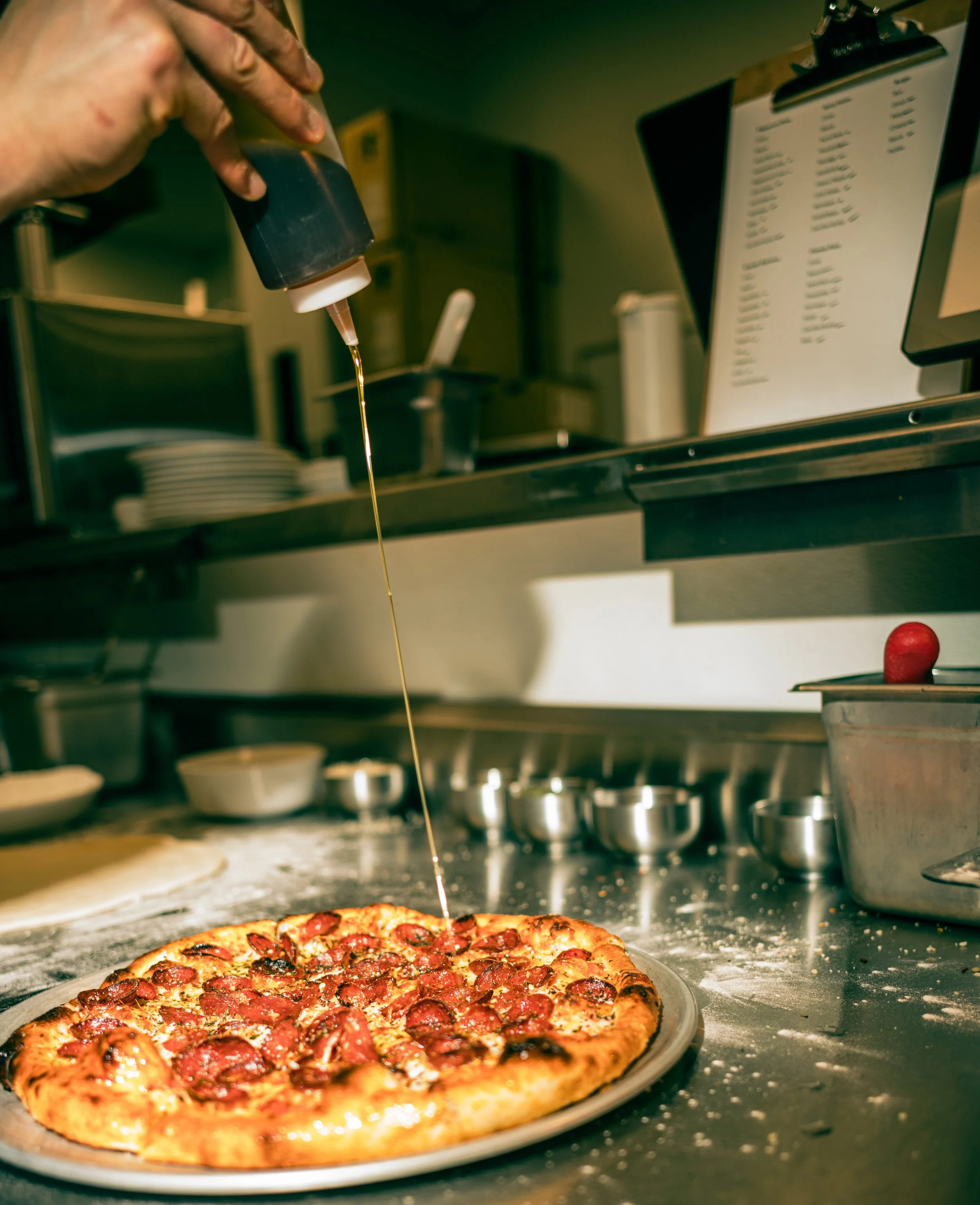 A pizza with pepperoni being topped with hot honey at a restaurant kitchen counter.