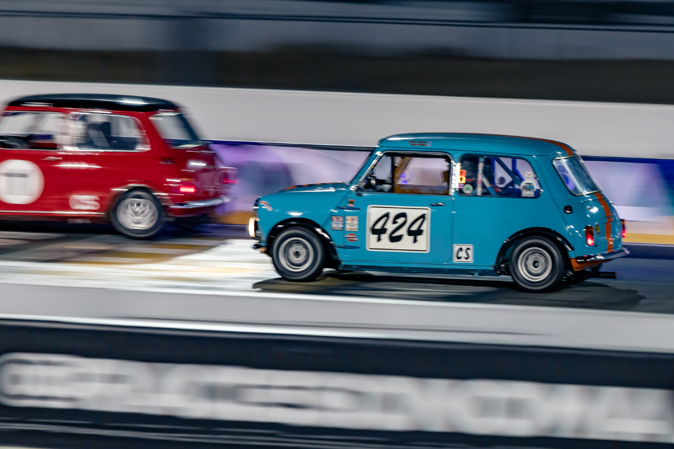 Two racing cars on a track, one blue Mini Cooper with the number 424 and one red vintage car, moving at high speed.