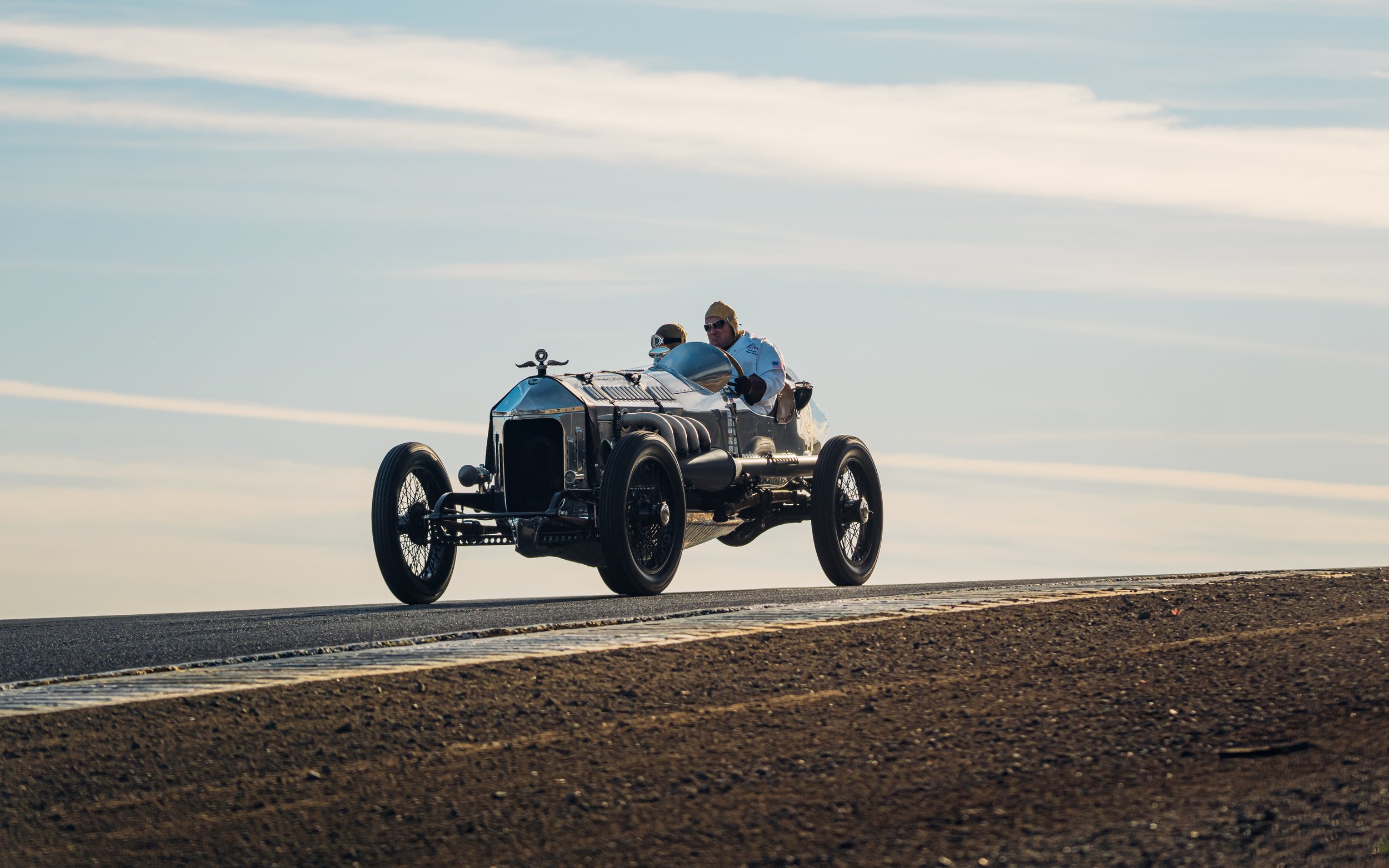 A vintage race car with two drivers on an open track during the daytime with a blue sky and some clouds.