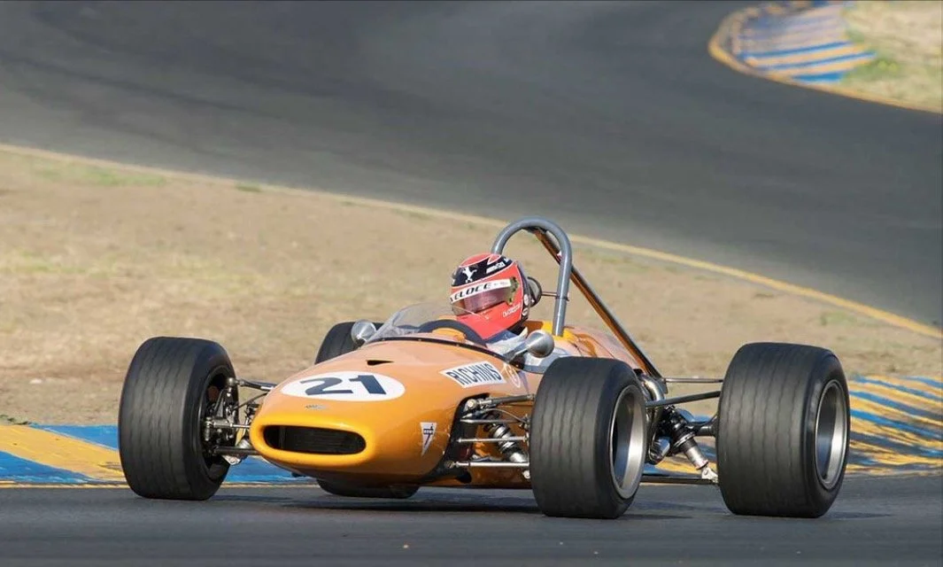 A vintage orange Formula race car with the number 21 on the front, driven by a person wearing a red helmet, speeding on a racetrack with a curved section and yellow and blue track markings.