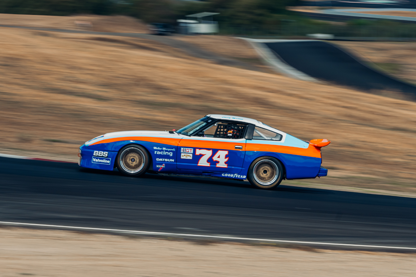 A vintage race car number 74 with a blue, orange, and white paint job racing on a track with a blurred background.