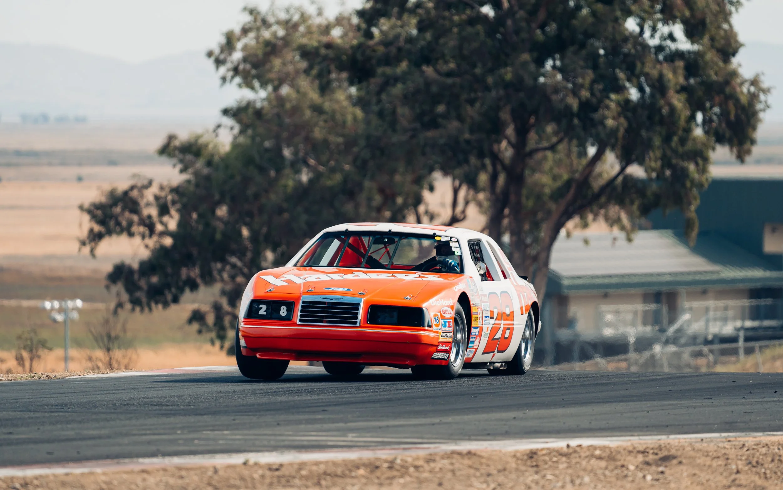 A vintage orange and white stock car racing on a track with a tree and a small building in the background.
