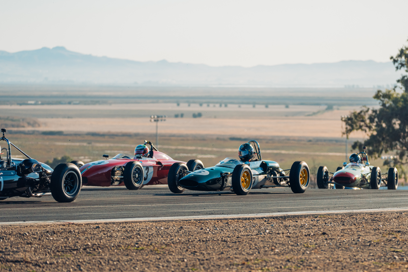 Three vintage Formula racing cars on a racetrack, with hills in the background and a tree on the right side.