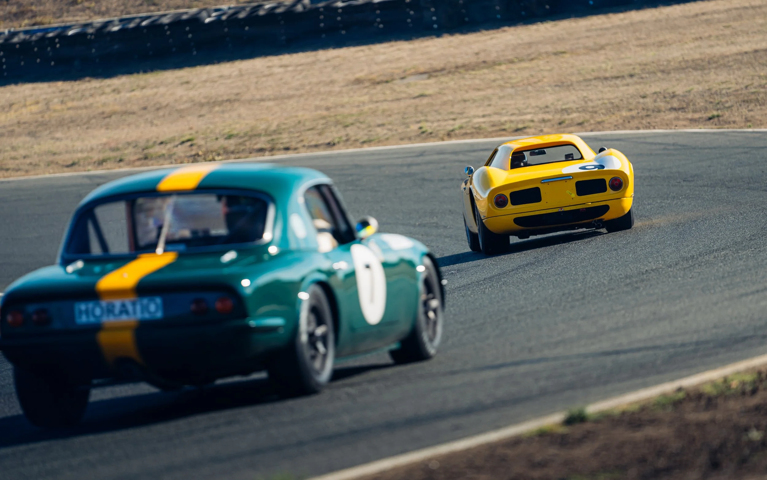 Two vintage race cars, one green with a yellow stripe and the number 0, and the other yellow with a rear license plate reading 'HORATIO,' racing on a track.