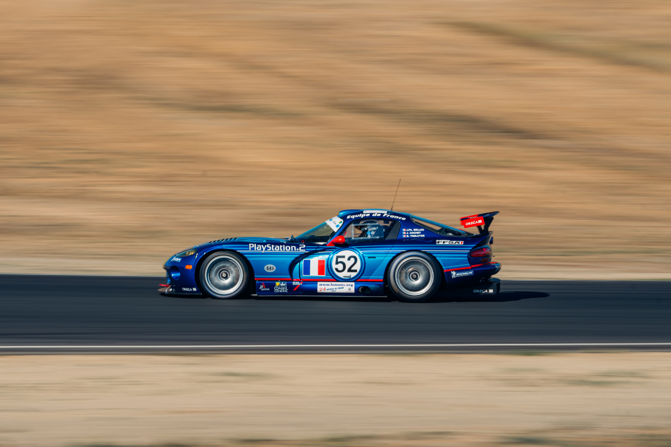 A blue race car with the number 52 on it speeding on a racetrack with a blurred background.