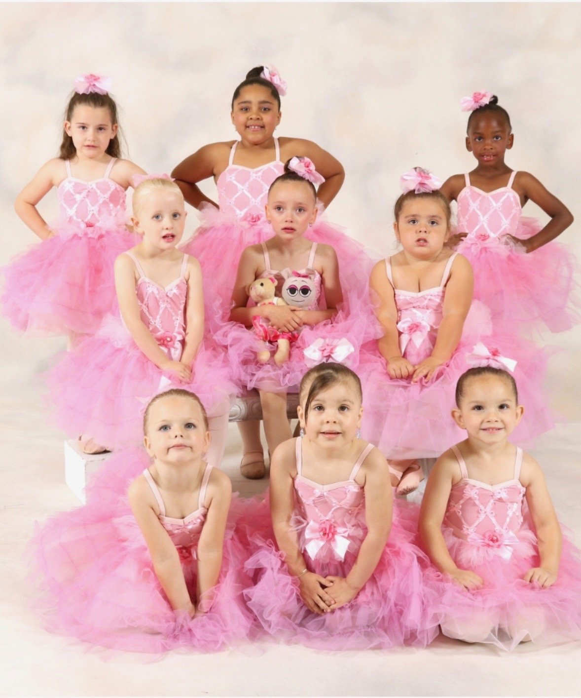 Group of young girls dressed in pink ballet costumes with tulle skirts and bows, sitting and standing for a photo.