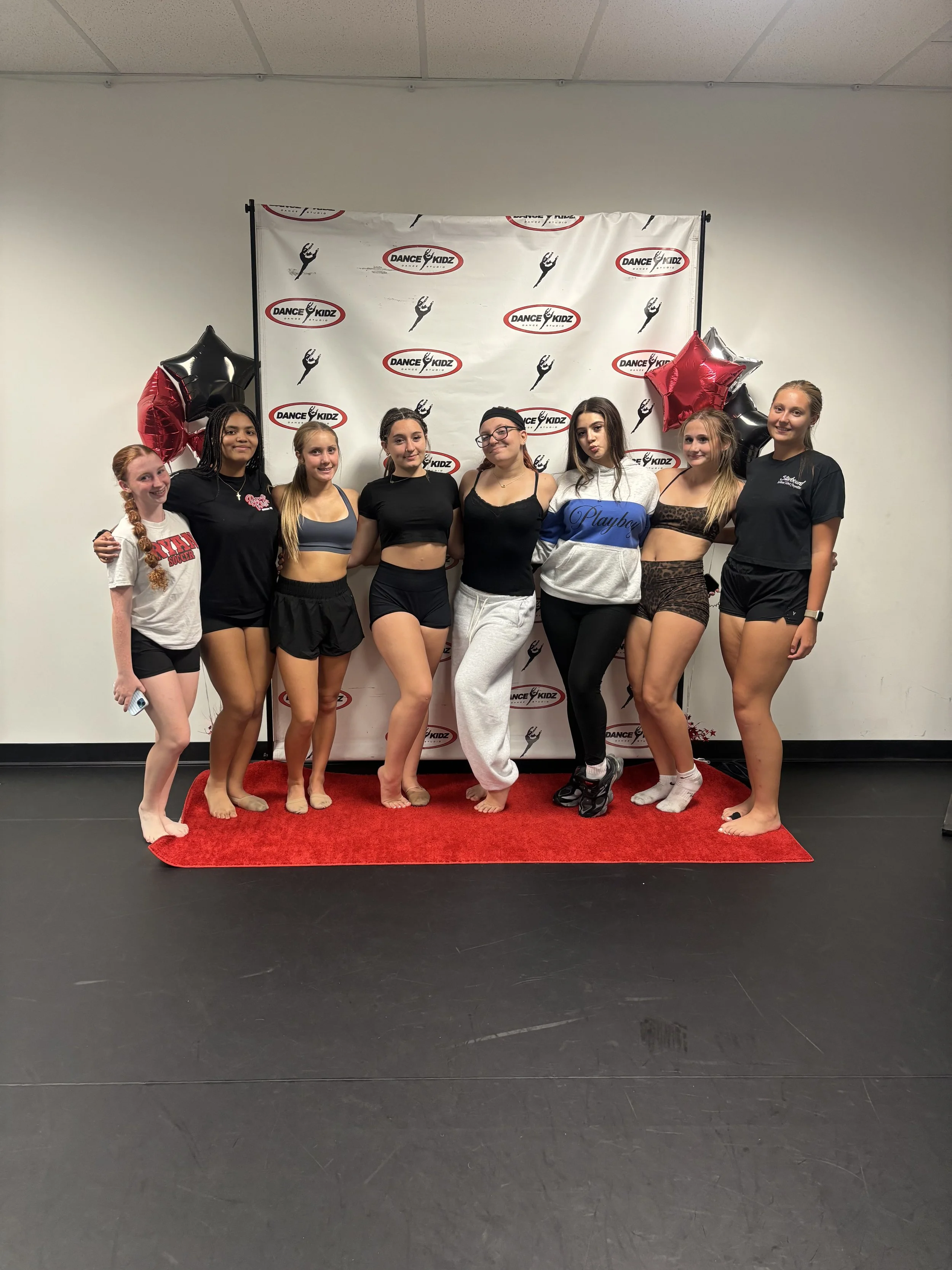 Group of nine young women posing for a photo in front of a backdrop with 'Dance Kidz' logos, celebrating a dance event. They are standing on a red carpet, wearing dance or casual attire, with balloons on either side.