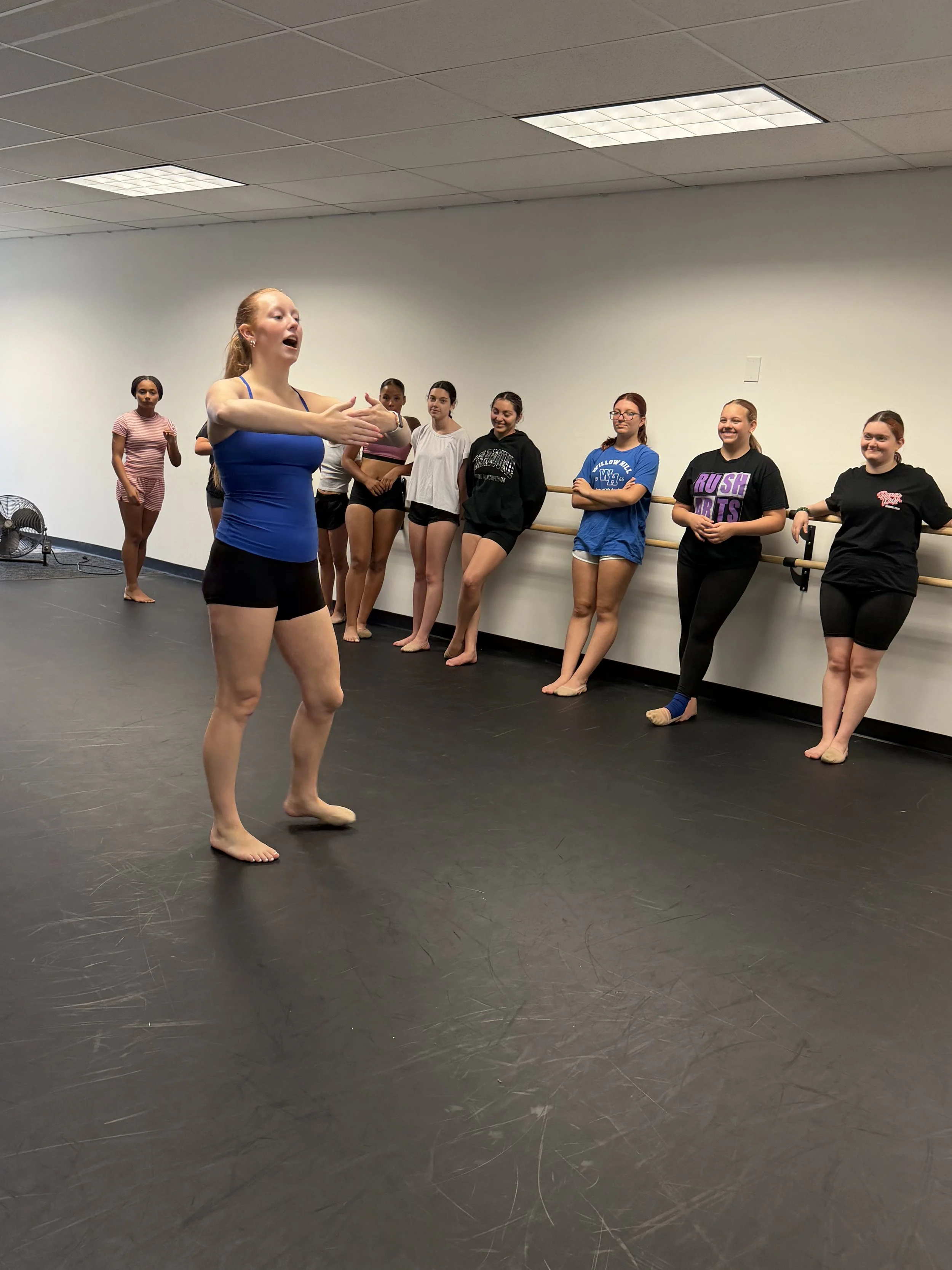 A ballet or dance class is taking place in a studio with a black floor and a white wall. Multiple young girls are standing near the ballet barre, dressed in casual dancewear or hoodie and shorts. An instructor or dancer is in the foreground, dressed 