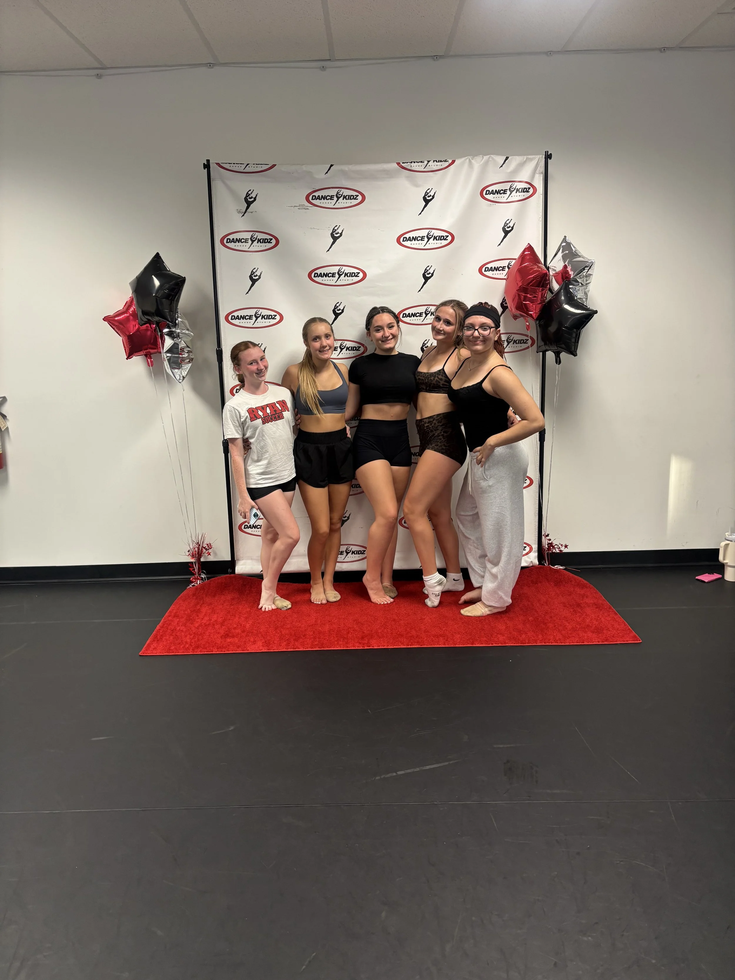 Five young women standing on a red carpet in front of a backdrop with 'Dance Kidz' logos, surrounded by red, black, and silver balloons, in an indoor dance studio.
