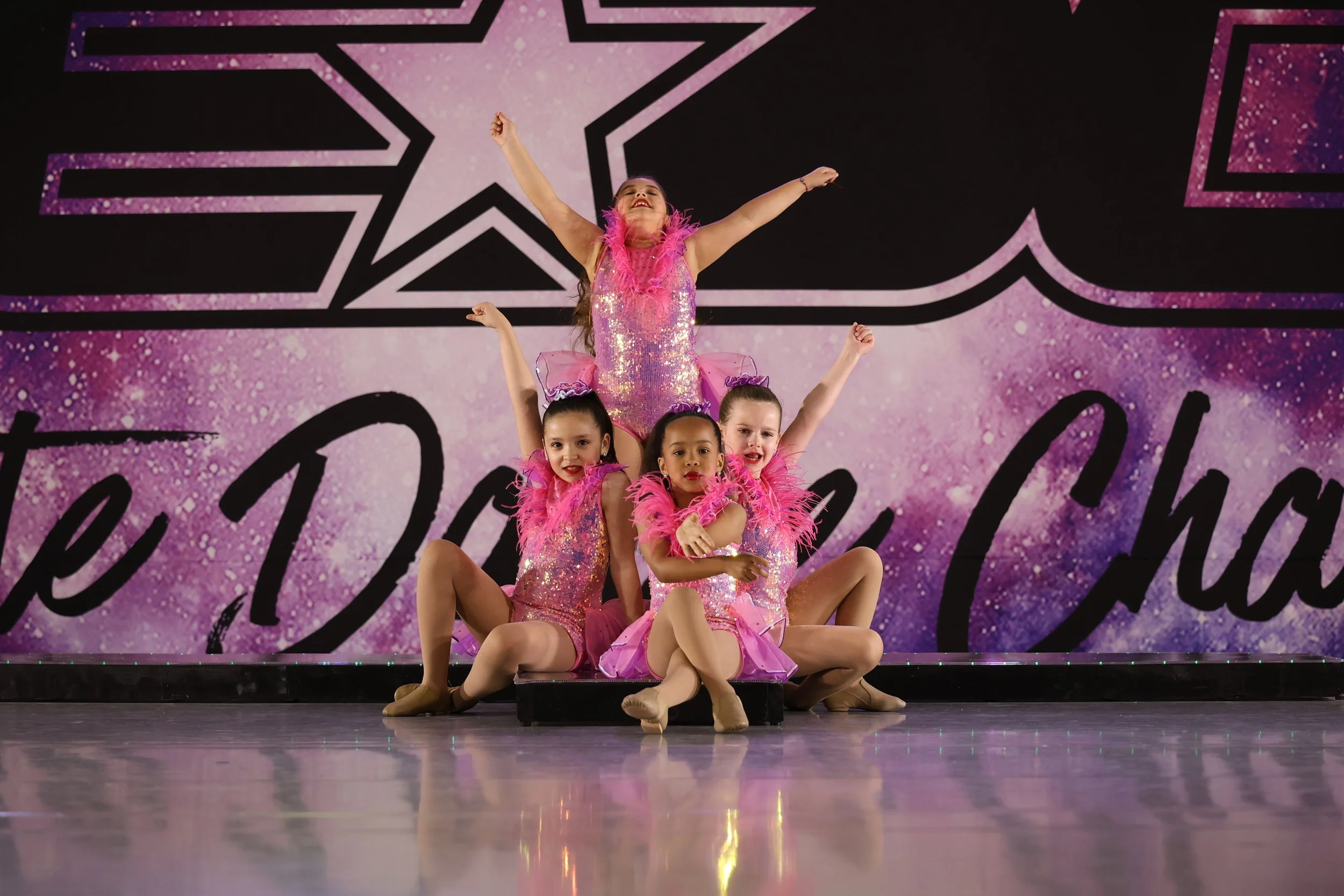 Group of young girls in pink and purple costumes performing a dance routine on stage with a star and musical note backdrop.
