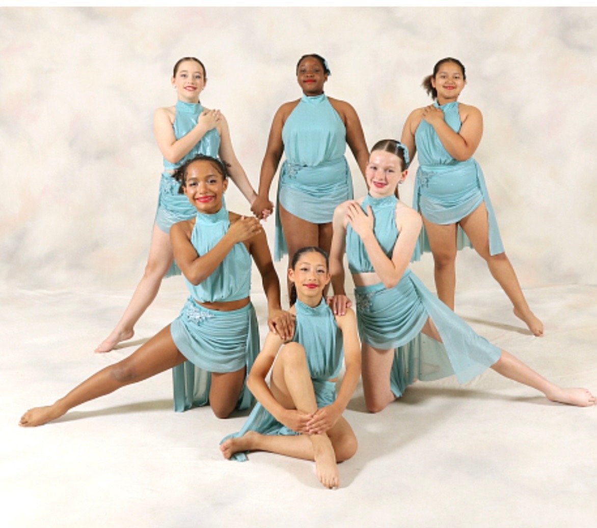 Seven young girls in matching light blue dance costumes pose together in a studio, smiling and holding hands.