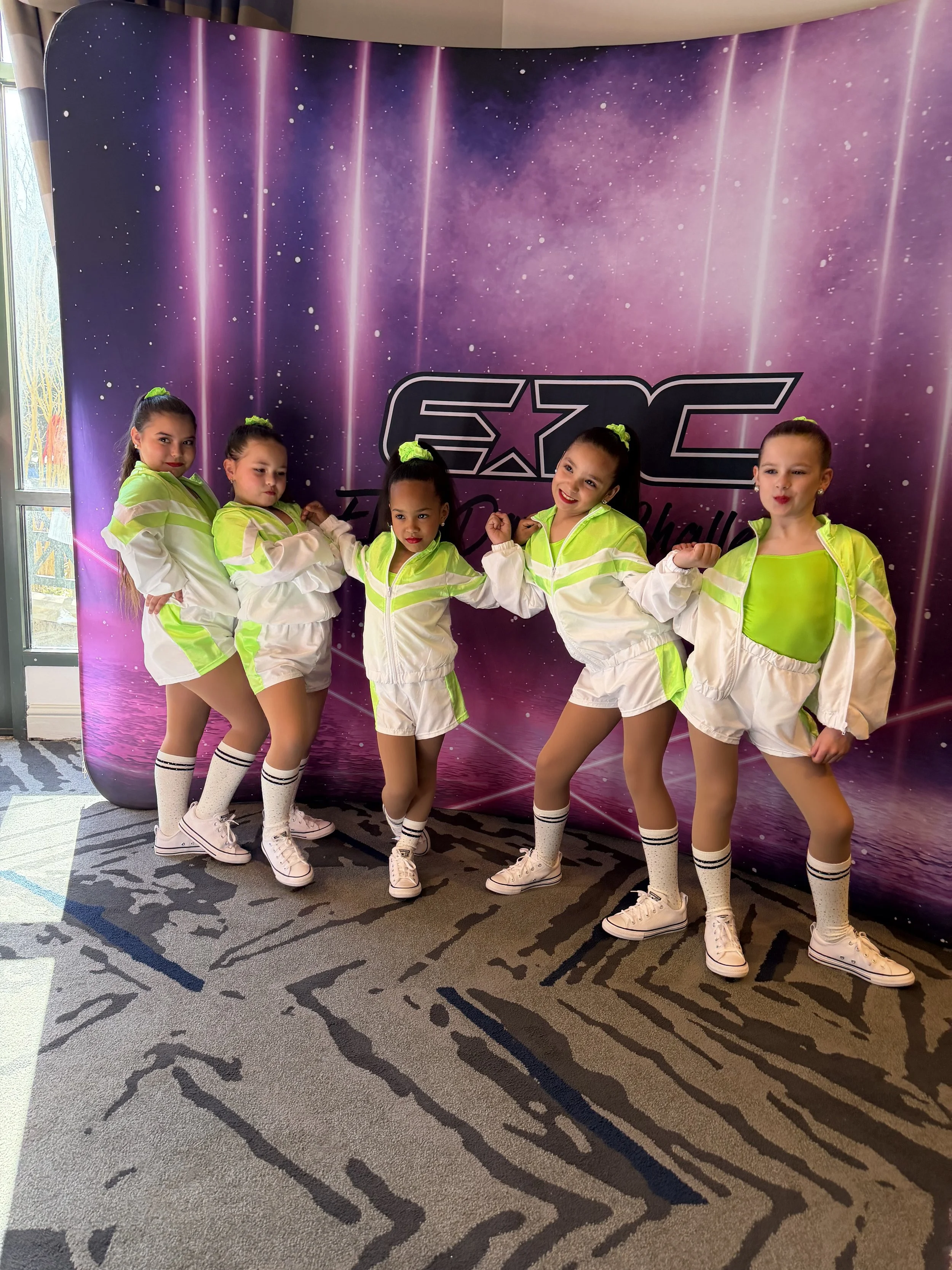 Six young girls dressed in matching neon yellow and white dance costumes with high ponytails and sneakers pose in front of a purple Galaxy-themed backdrop.