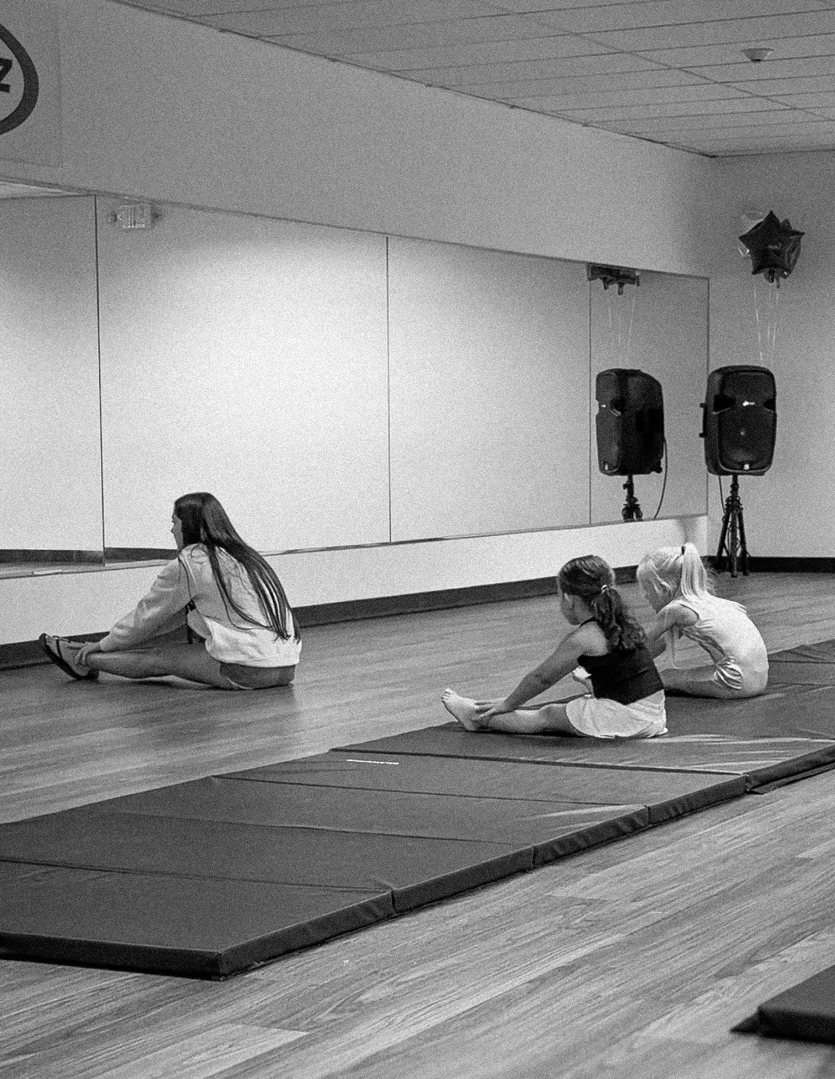 Three young girls sitting on exercise mats in a dance or yoga studio, stretching their legs forward. Two girls are facing away from the mirror on the wall, while one girl is facing towards the mirror. There are speakers and a no smoking sign on the wall in the background.