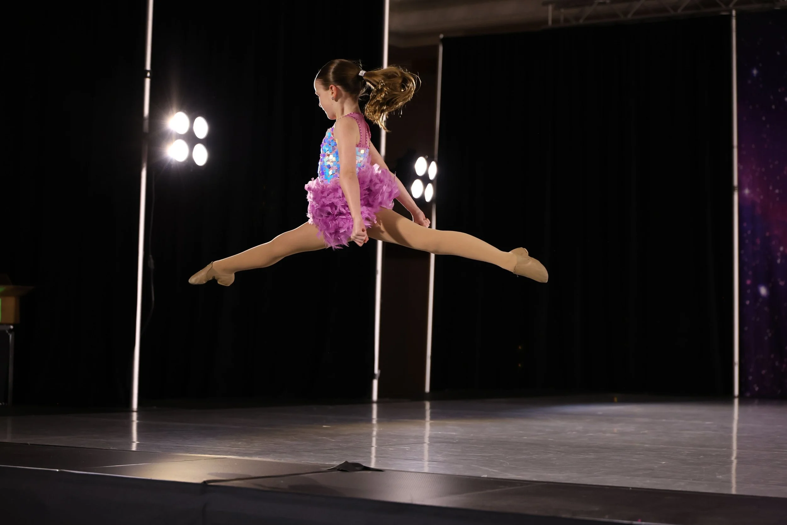 A young girl in a colorful dance costume performing a split on stage during a dance competition.