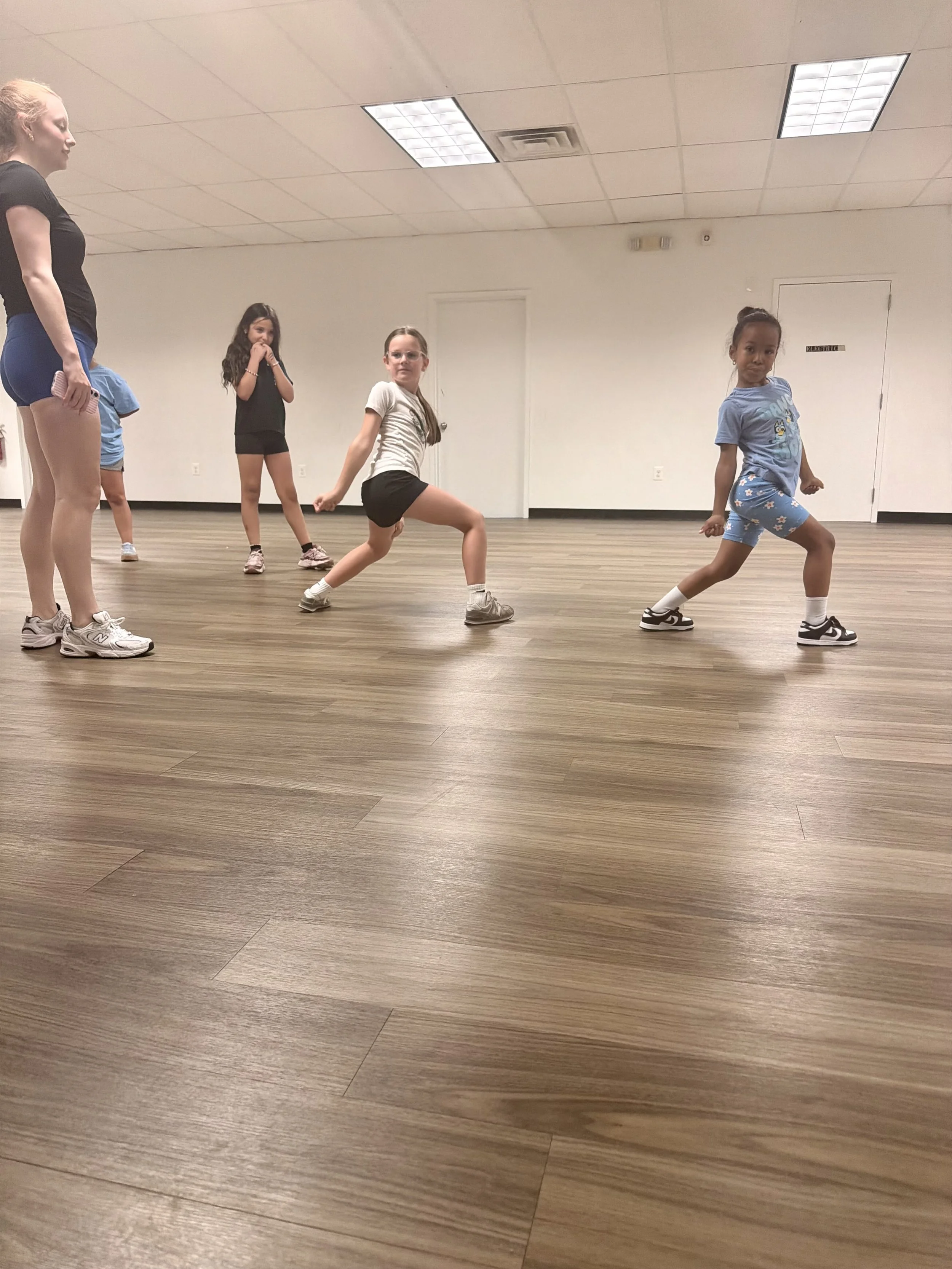 A group of young girls dancing in a spacious room with a wood floor and white walls, under a ceiling with fluorescent lights.
