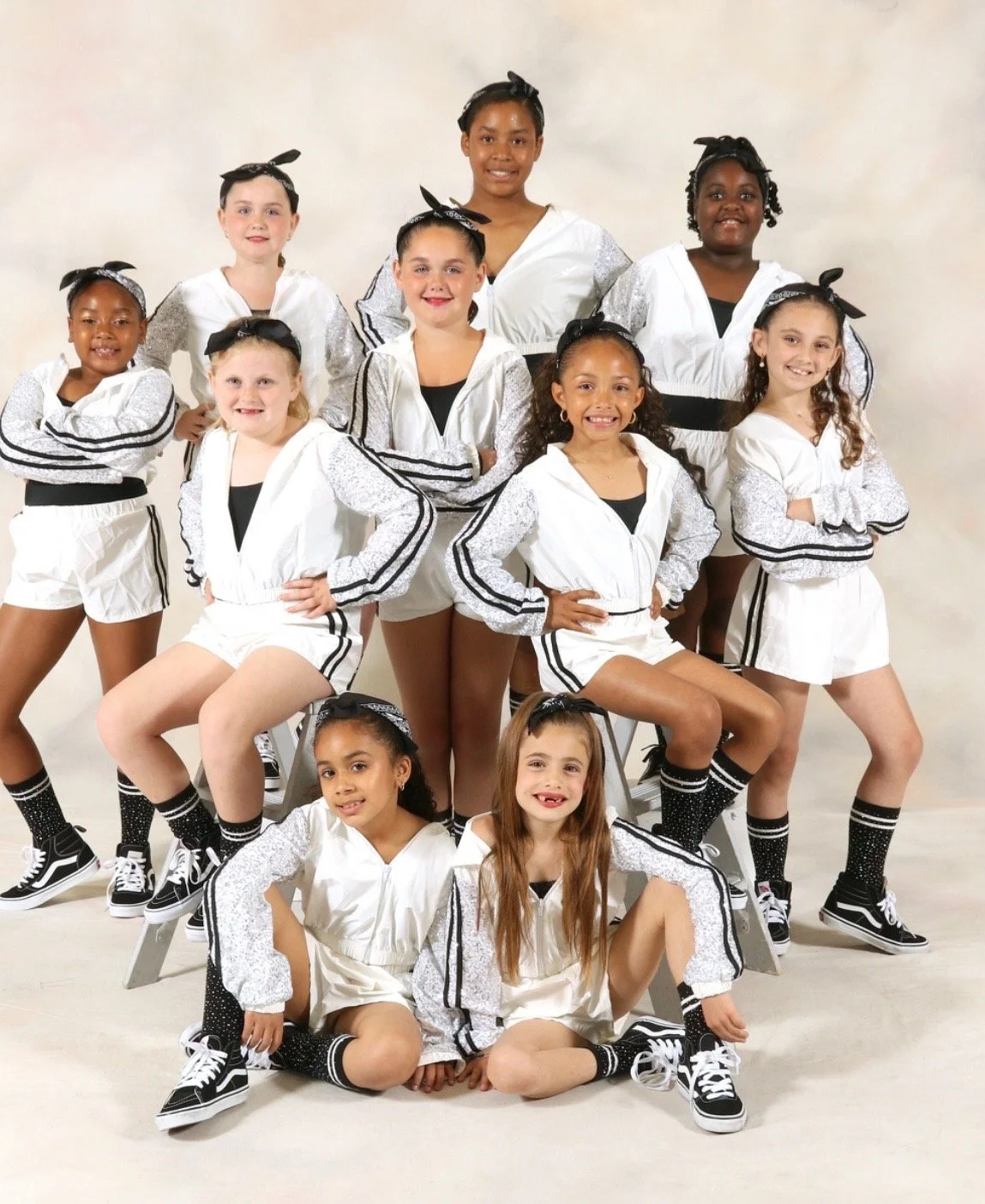 A group of ten young girls in matching white and black dance costumes, posing together for a studio photo against a neutral background.