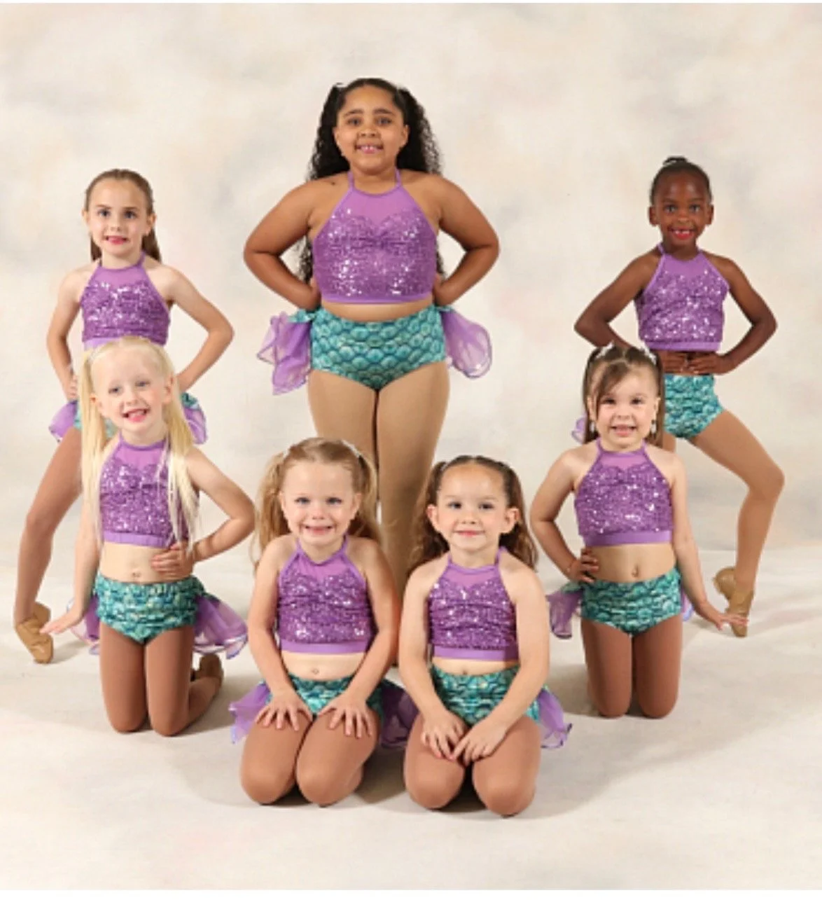 Group of seven young girls in matching purple sequined costumes with teal shorts and purple sashes, posing for a dance performance in front of a plain backdrop.
