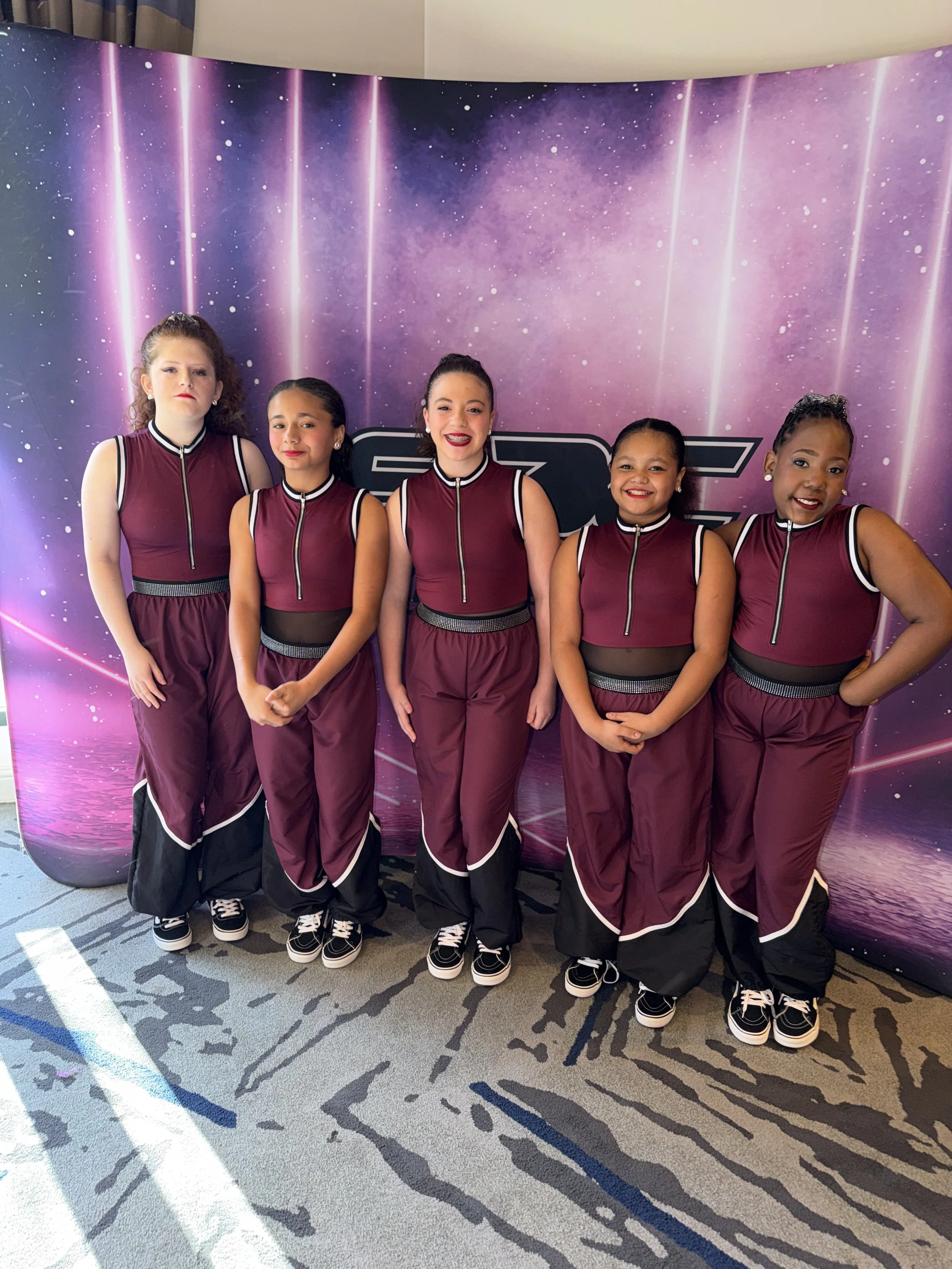 Five young girls in matching maroon and black dance costumes standing in front of a purple cosmic-themed backdrop. They are inside a room with patterned carpet and light coming from the left side.