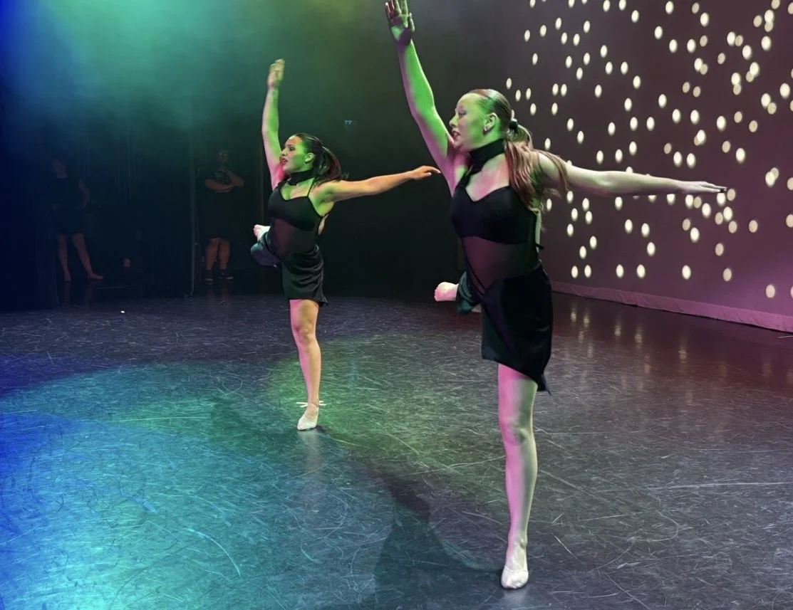Two female dancers in black outfits with mesh midsections perform a dance routine on stage with their arms raised and one leg lifted. The stage is dark with green and purple lighting, and a backdrop with white dot patterns.