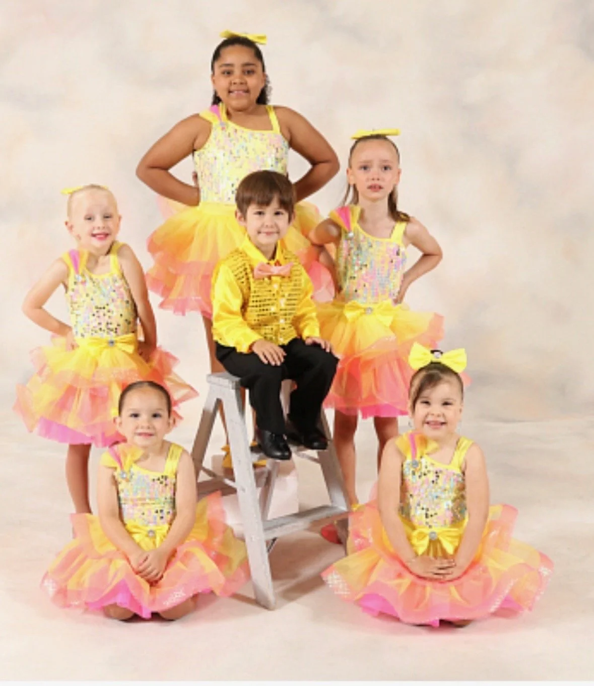 A group of young children, mostly girls, dressed in colorful yellow and pink tutus and top costumes, with one boy in a yellow jacket and black pants, posing for a photo in a studio with a plain background.