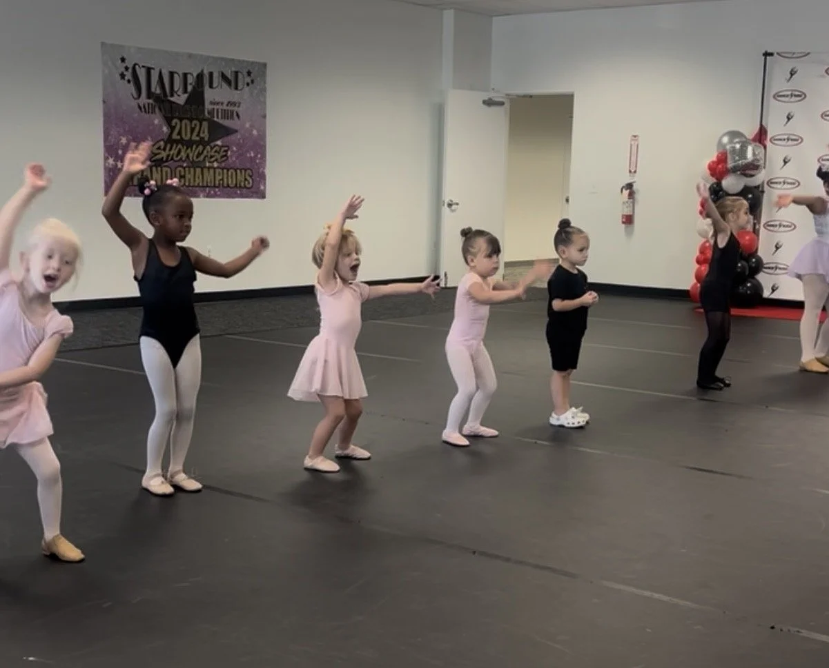 Young girls in ballet outfits practicing dance moves in a studio with a banner for a 2024 recital.