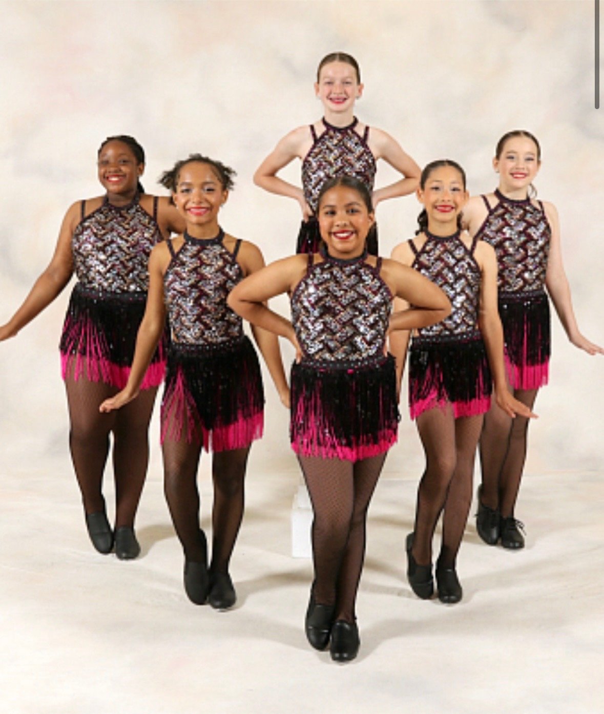 Six young girls in matching dance costumes posing in formation on a stage or studio. The costumes are black, pink, and patterned with shimmering details, and they are wearing black dance shoes and fishnet tights at dance kidz a dance studio Bensalem