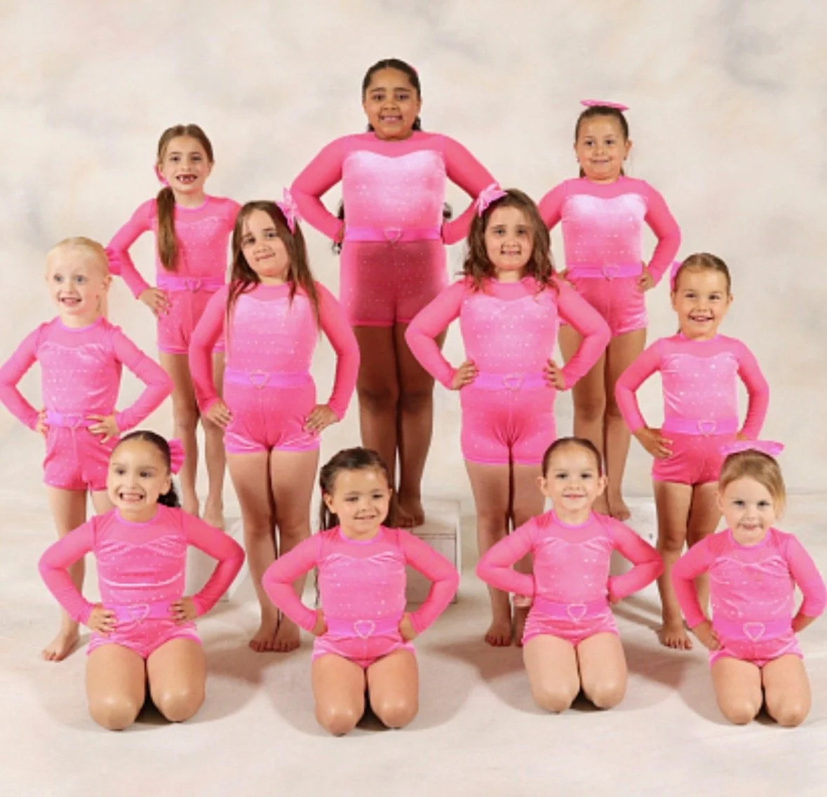 A group of young girls dressed in matching pink dance costumes posing for a photo, with some girls kneeling and others standing.