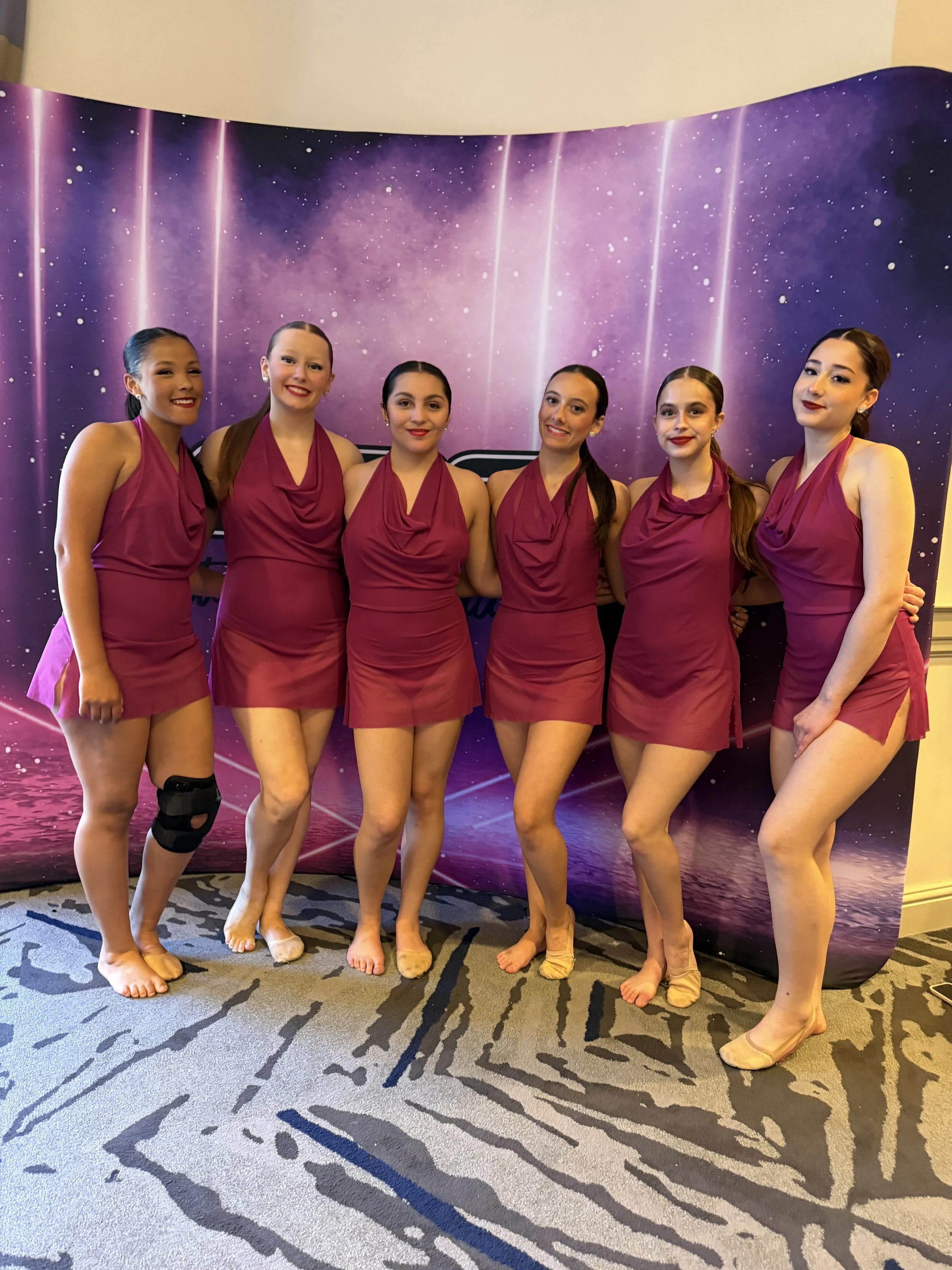 Six young women in elegant purple dresses, standing close together and smiling at the camera, backstage at a performance venue with a purple starry backdrop and patterned carpet.