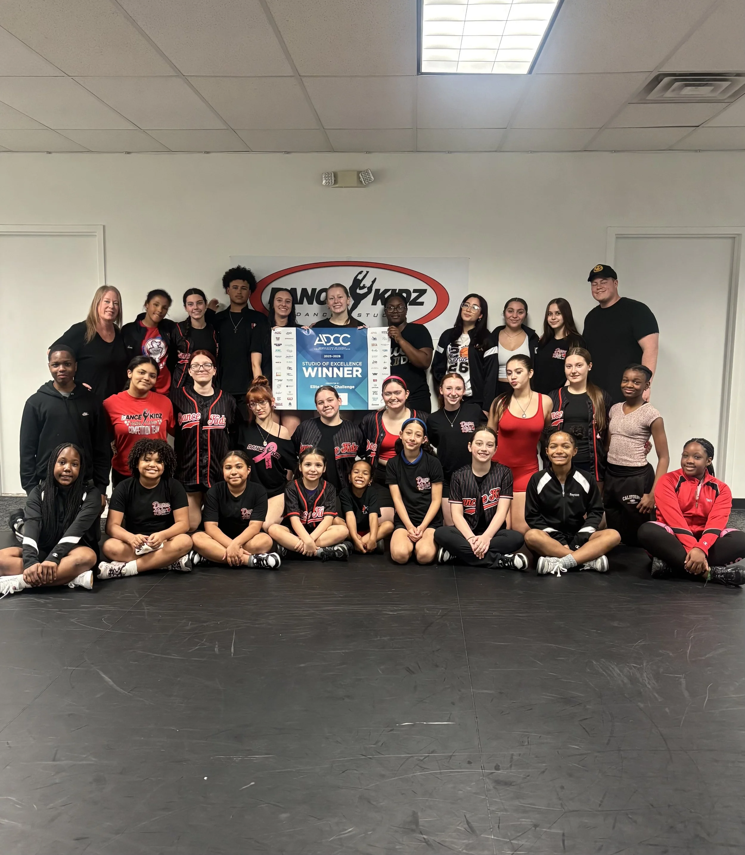 Group photo of children and adults at a dance studio, holding a large blue and white award certificate that reads "Studio of Excellence Winner" for the 2025-2026 season, in front of a "Dance Kidz" sign.