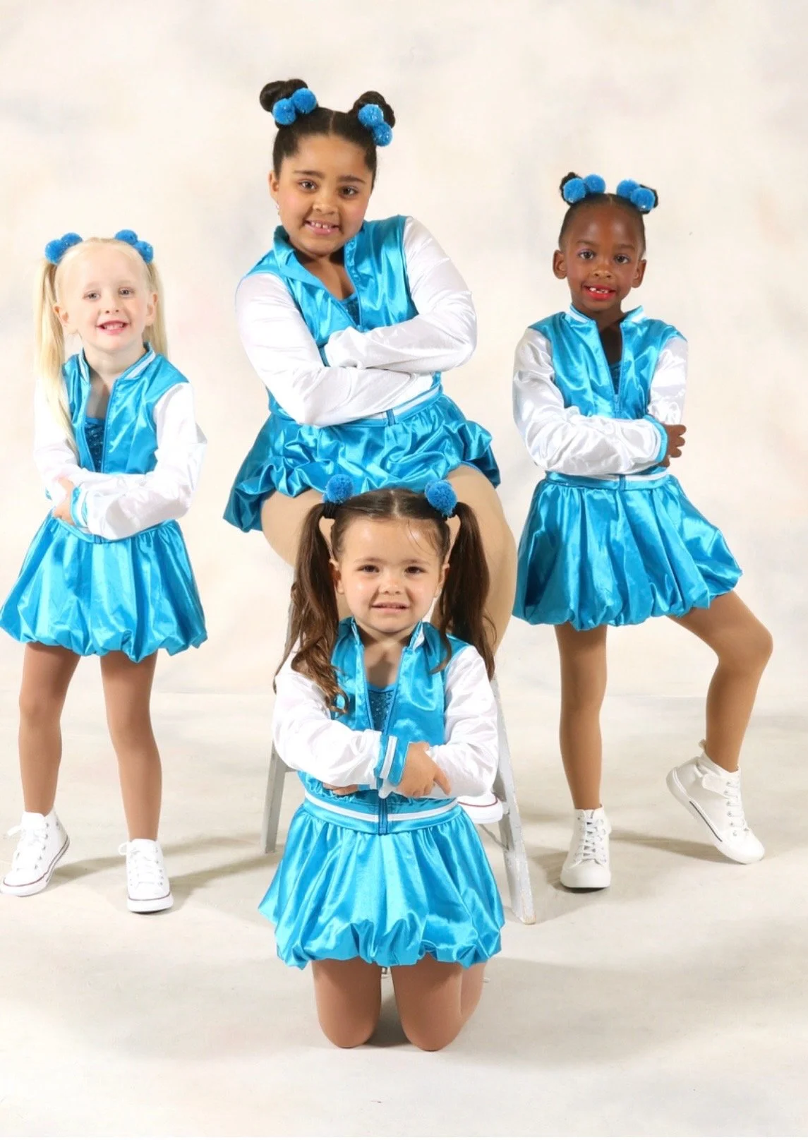 Five young girls dressed in blue and white cheerleading outfits with blue pom-poms in their hair. They are striking different poses against a plain white background.